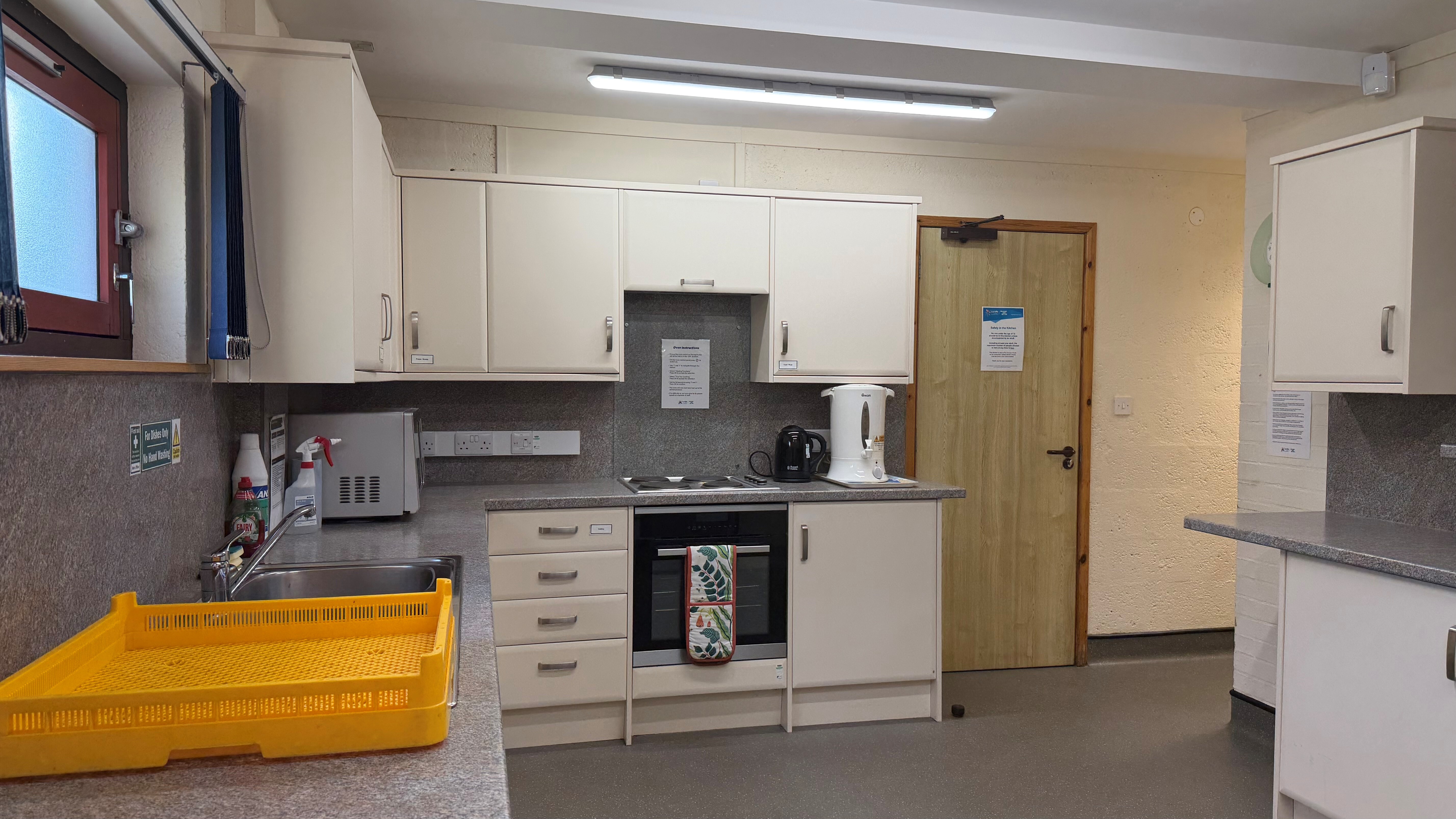 Kitchen area with electric hob, built-in oven, kettles, and cream cabinets beside a wooden door.