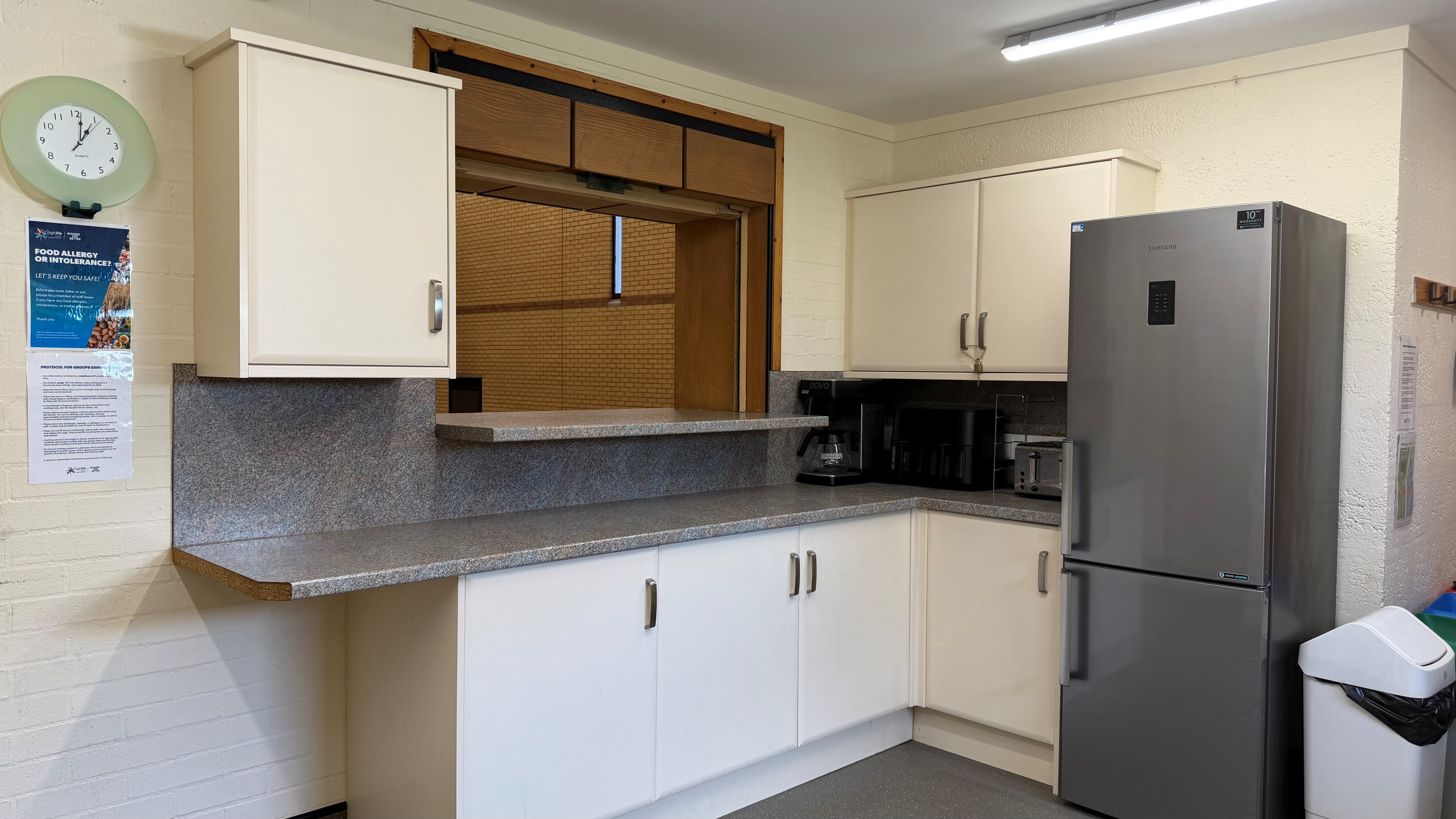 Kitchen serving hatch with coffee maker, toaster, fridge-freezer, and wall clock.