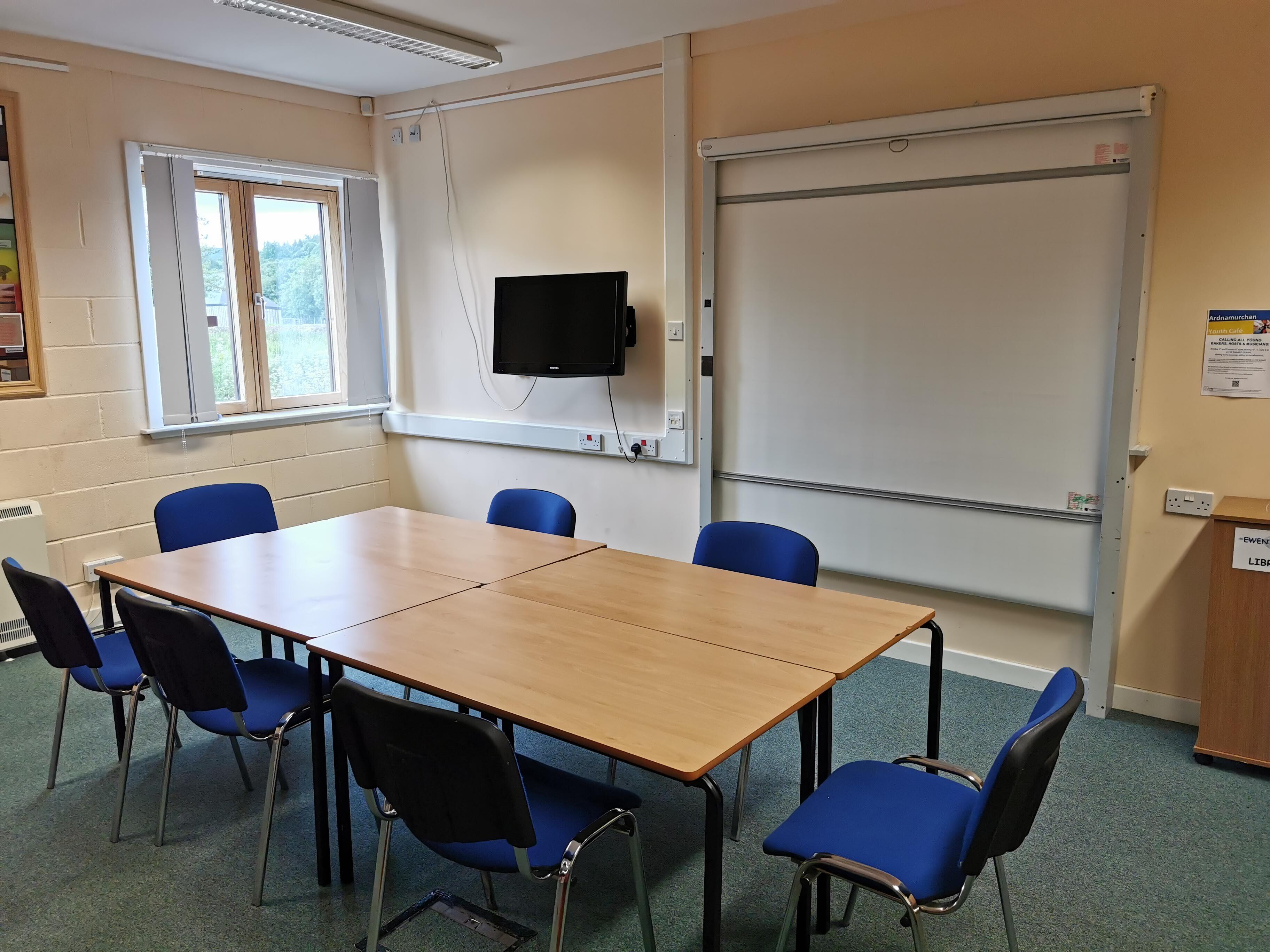 A meeting room featuring natural lighting, equipped with a central table surrounded by chairs, as well as a wall-mounted TV, a flip chart and a whiteboard.