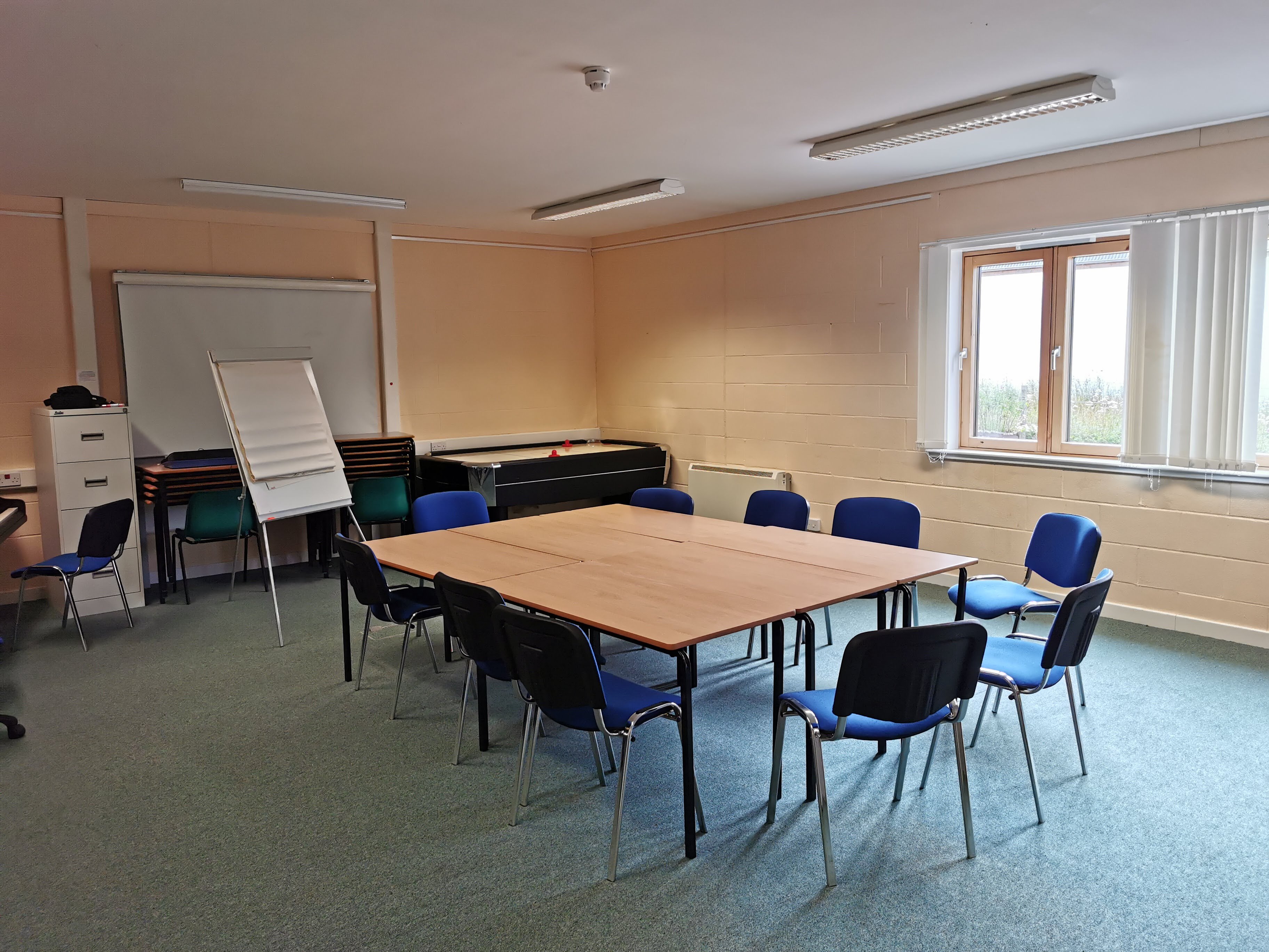 A meeting room featuring natural lighting, equipped with a central table surrounded by chairs, as well as a flip chart and a whiteboard.