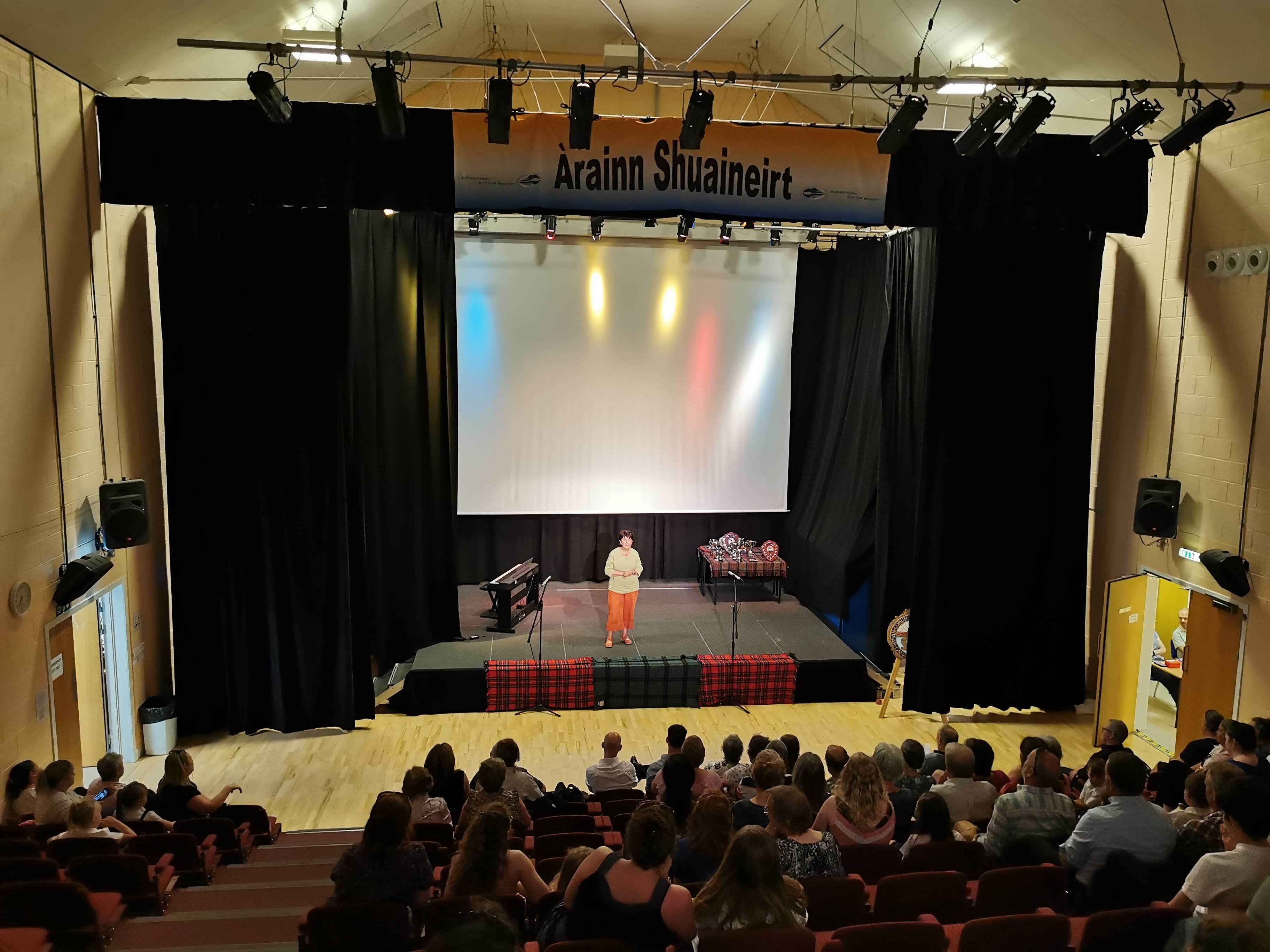 A theater featuring a central stage, equipped with a white screen, showcasing a solitary performer before an audience.