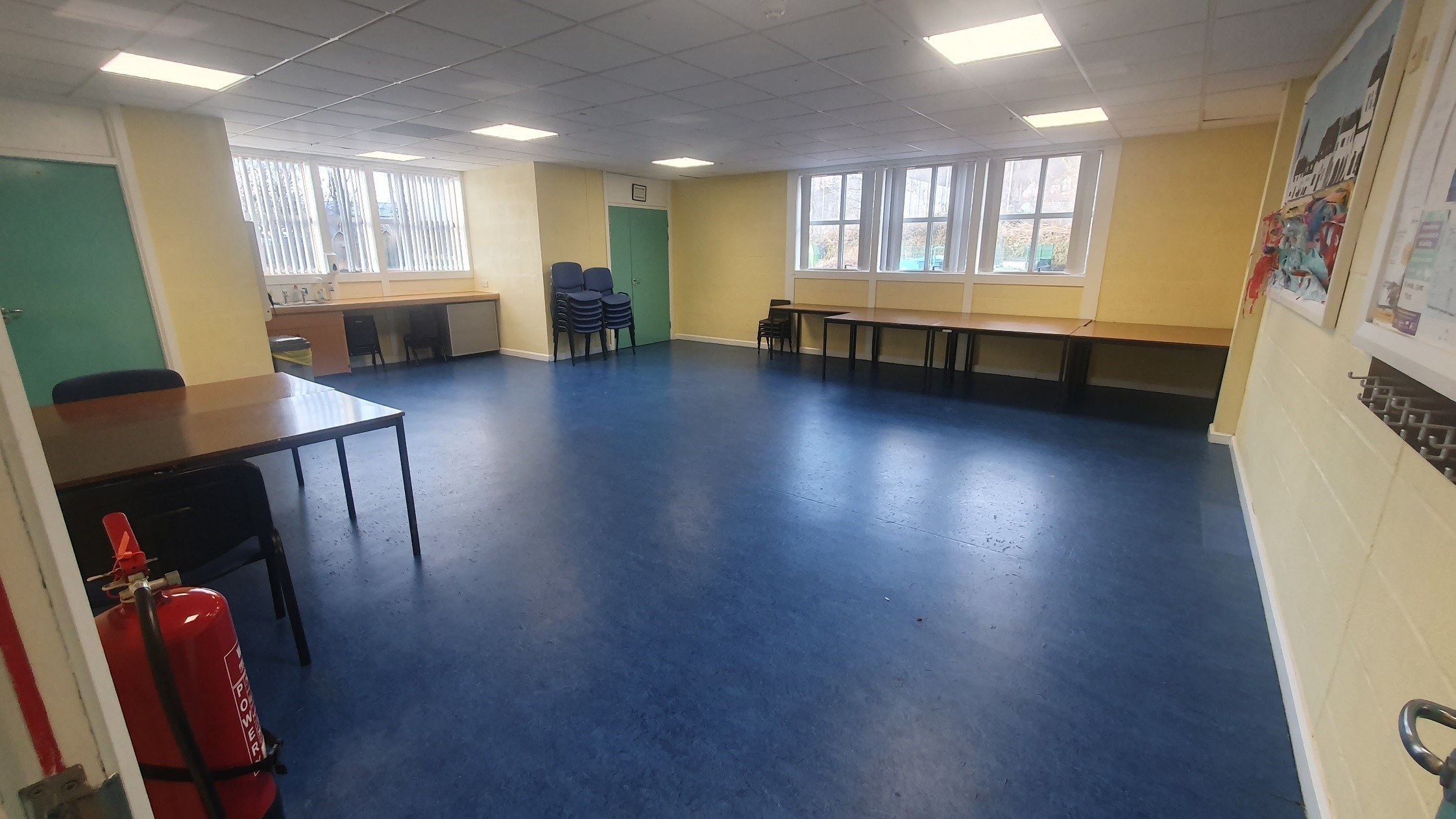 The Coigach room is a spacious area featuring laminate flooring and bright walls. Tables are arranged along the perimeter, complemented by stacked chairs that are readily available for seating.