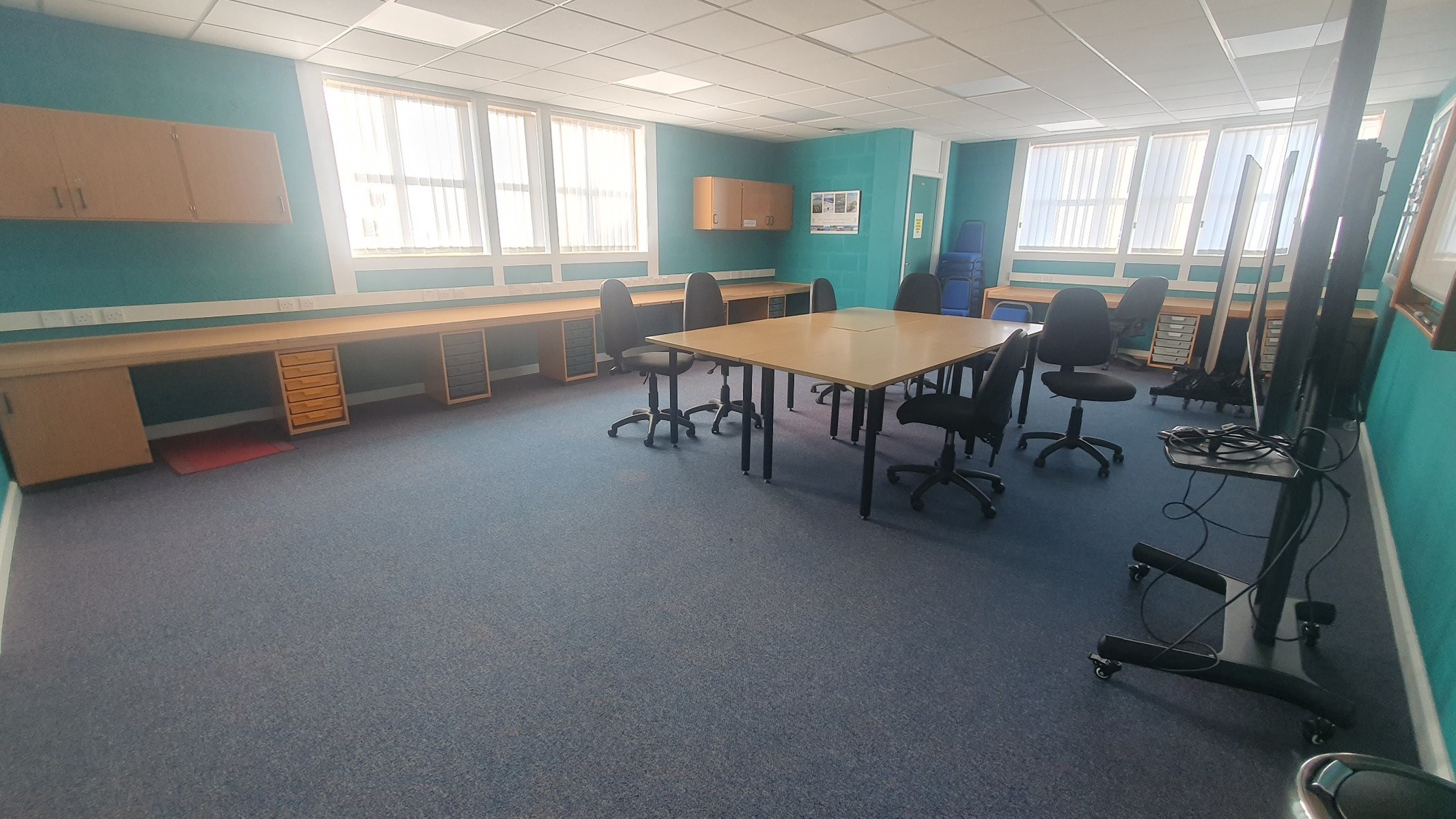 The Distance Learning Room is an expansive and well-lit space, featuring carpeting and centrally located large tables, which are surrounded by office chairs. Additional desk space is available around the perimeter of the room, enhancing its functionality.