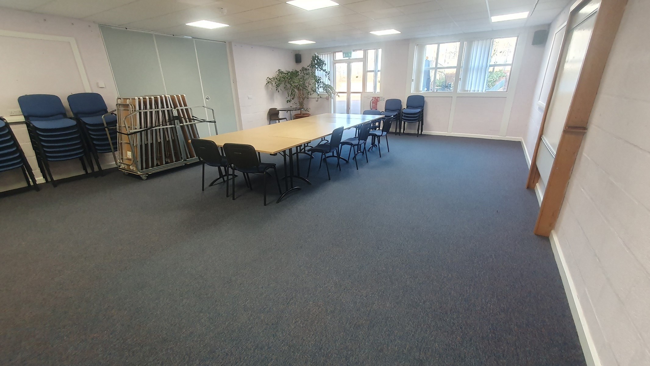 The Lochbroom room is a spacious, carpeted area illuminated by natural light streaming through its windows and a fire door. Centrally positioned tables are accompanied by surrounding chairs. Additionally, a dividing door connects this room to the Assynt room.