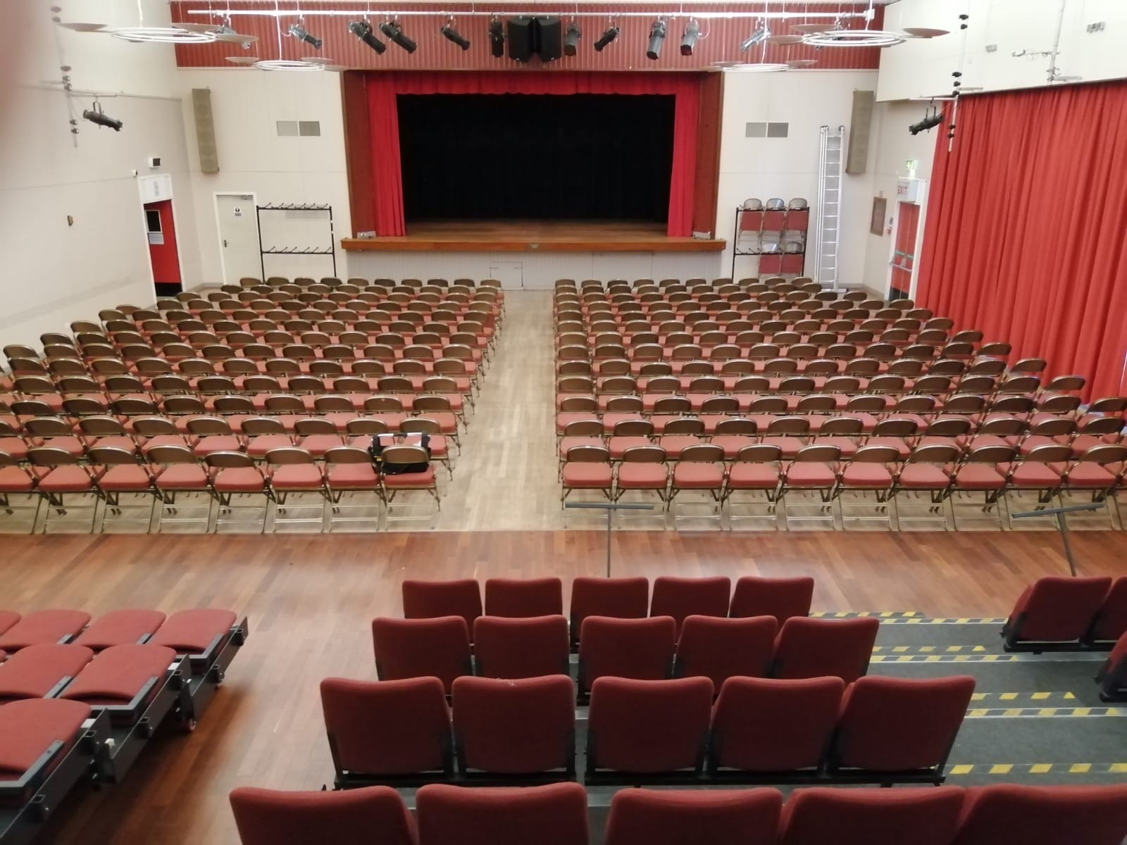 A hall featuring a central stage. There is retractable seating at the back of the room, along with additional seating arranged in front of the stage.