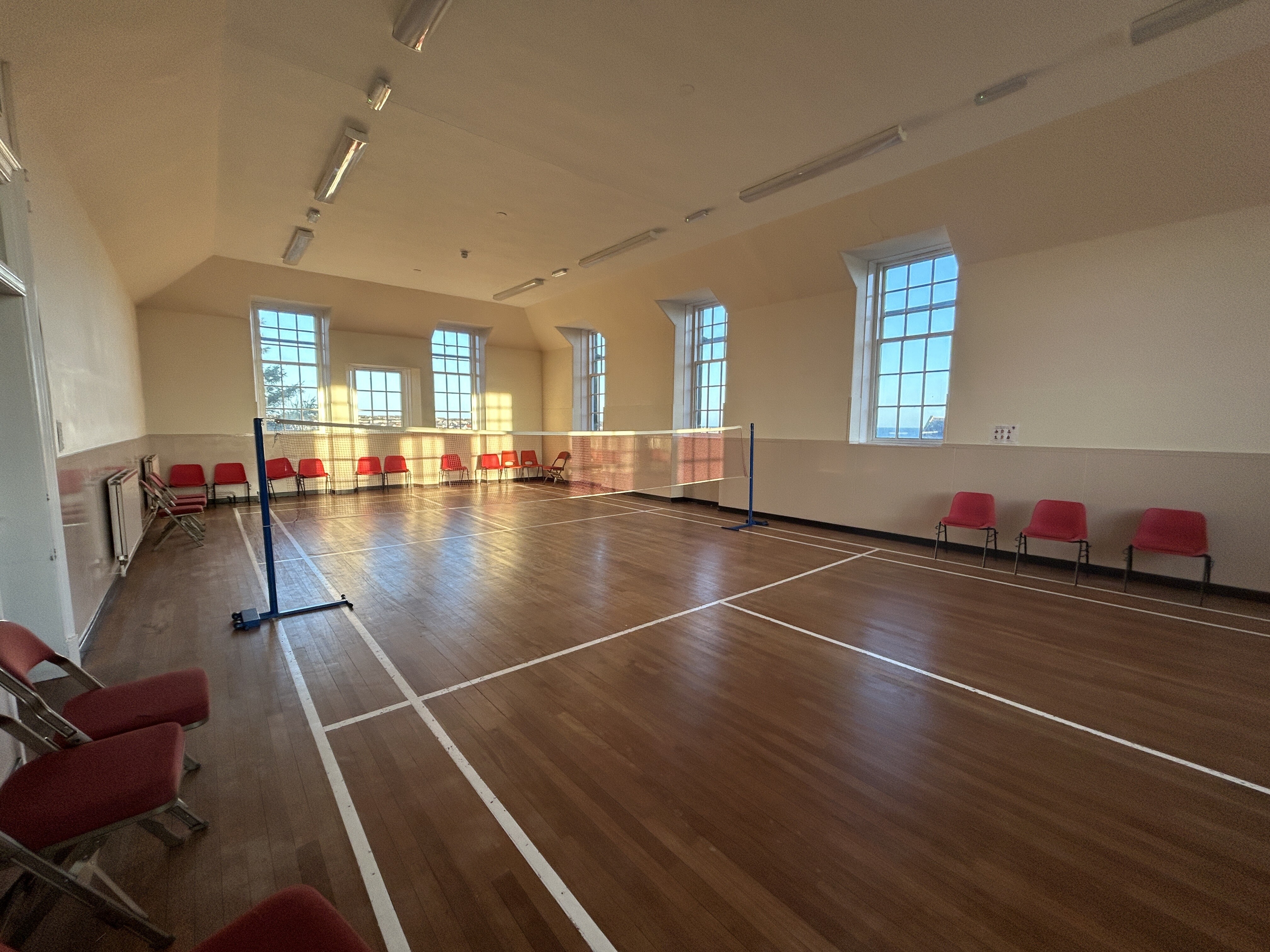 A well-lit hall featuring a badminton court complete with a net, with chairs arranged along the sides of each wall.