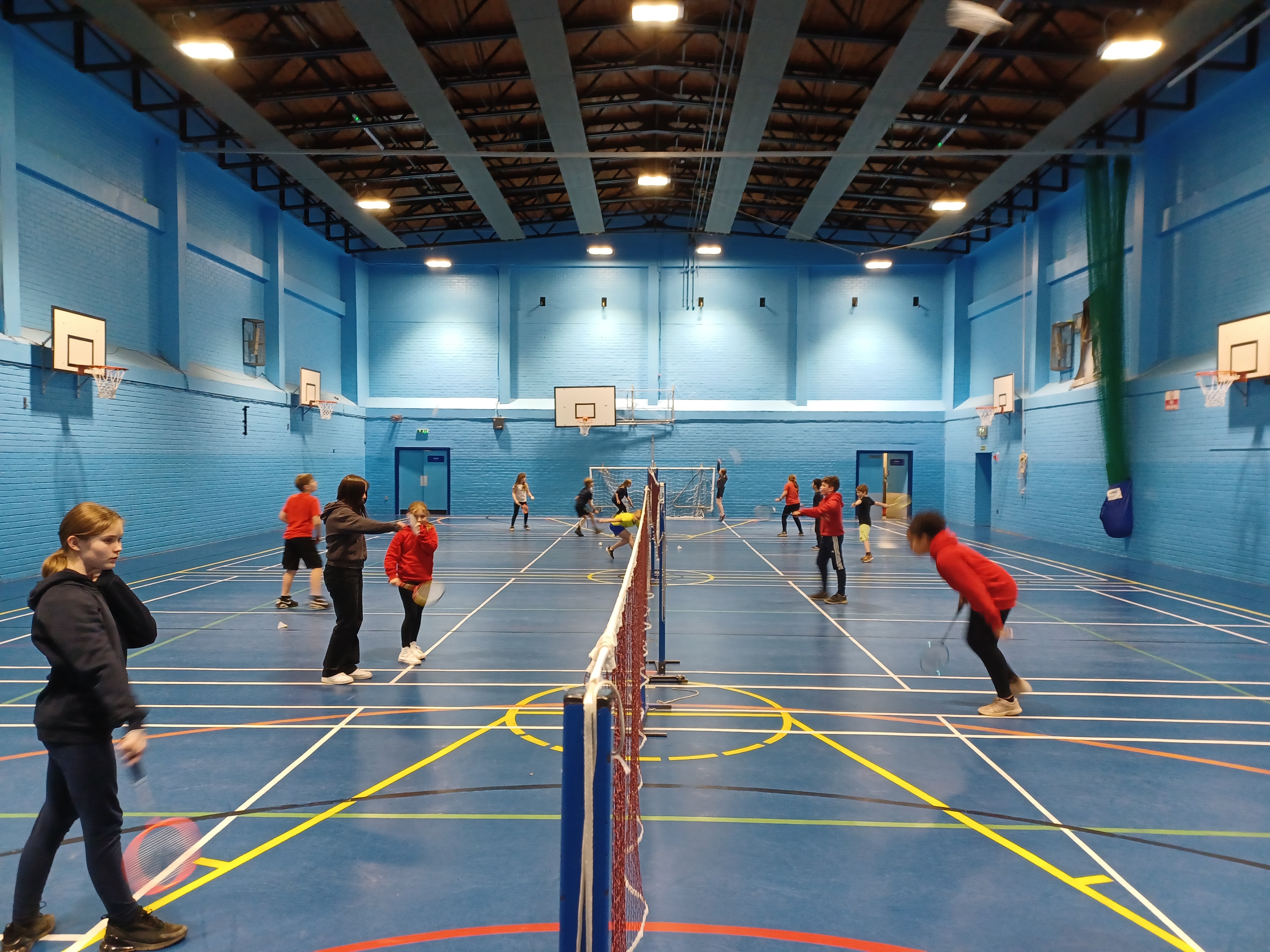 Children playing badminton across all courts of a large gym hall.