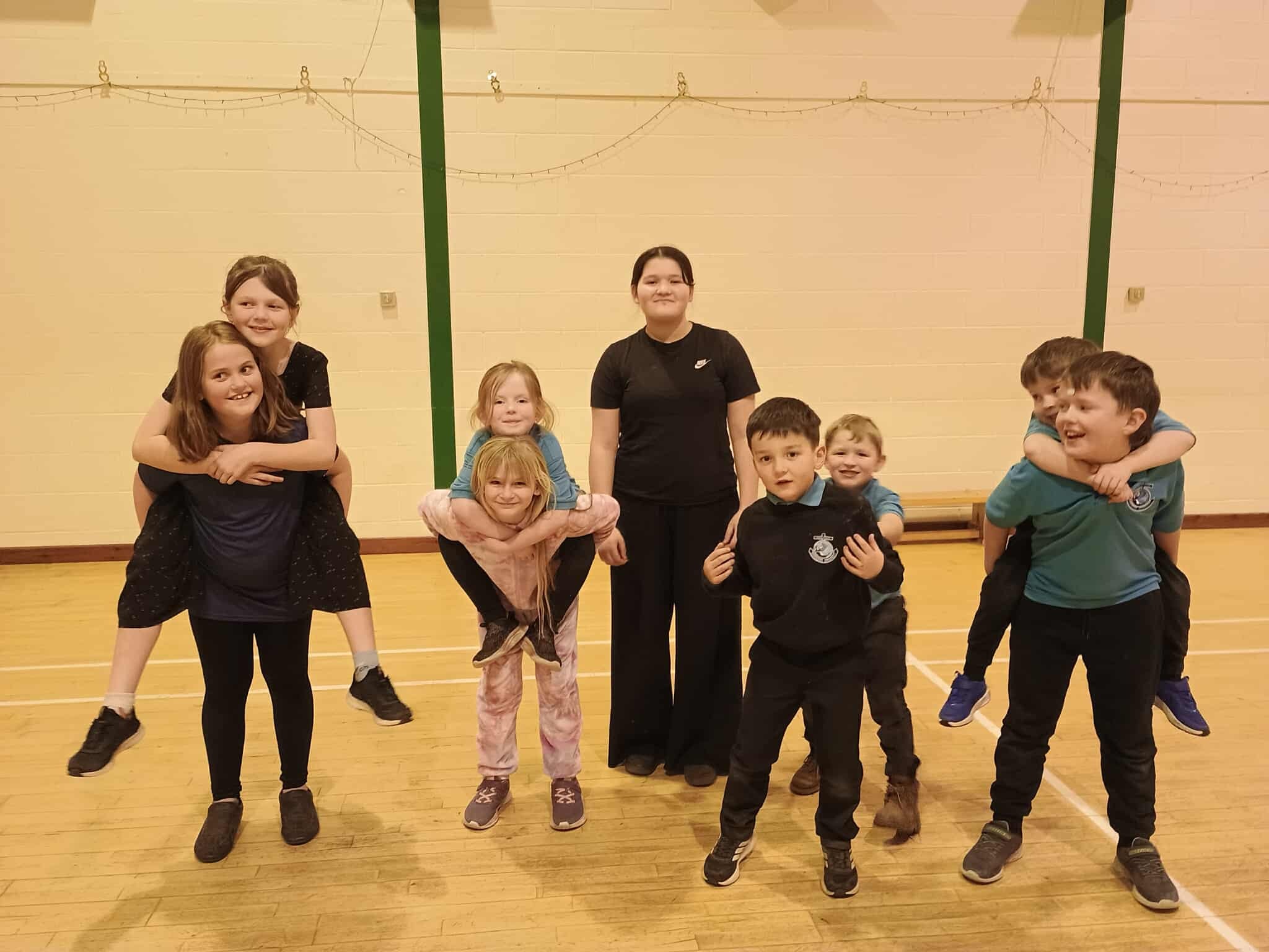 A group of smiling primary school children in a gym hall giving piggybacks.
