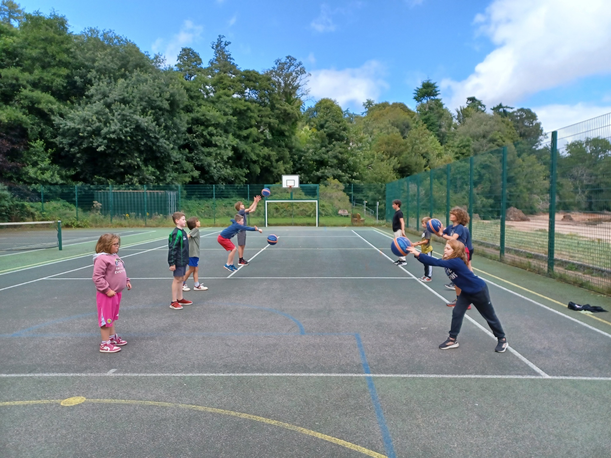 A group of primary children on an outdoor basketball court practicing chest passes with basketballs.