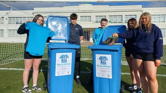 A group of secondary Young Leaders standing around two blue wheelie bins on a pitch, and one putting a pair of secondhand football boots into one of the bins.
