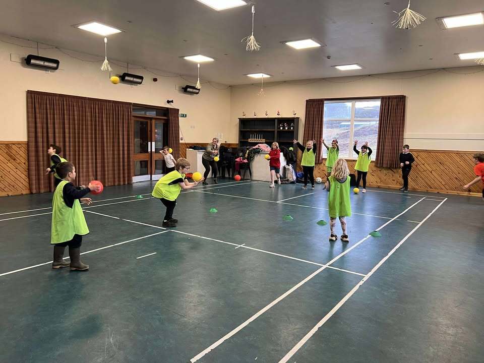 Young children in a gym hall in Kinlochbervie engaged in ball throwing activity.