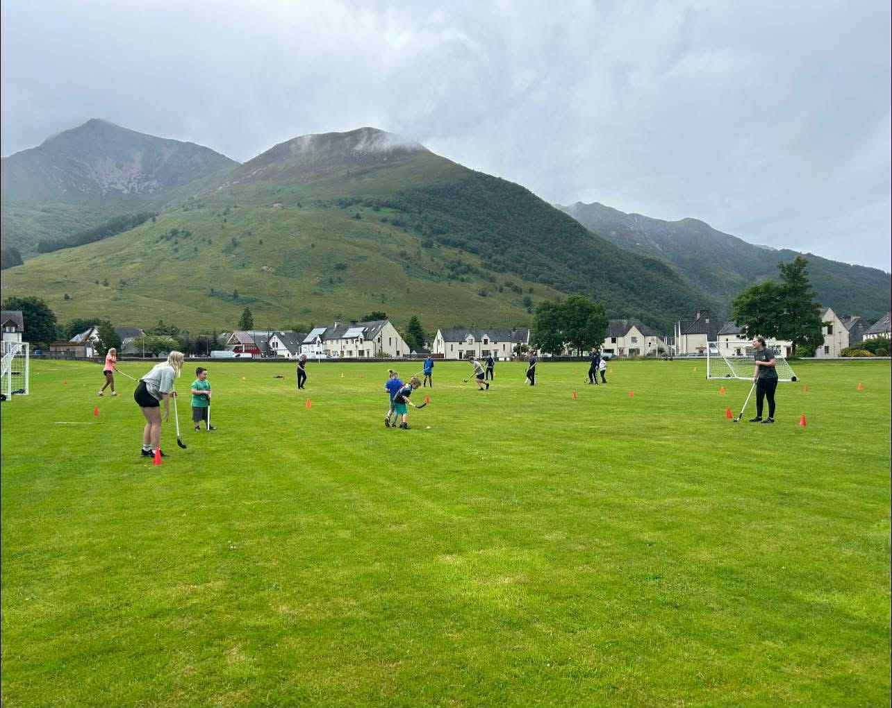 Children scattered across a pitch in Lochaber playing shinty with mountains in the background.