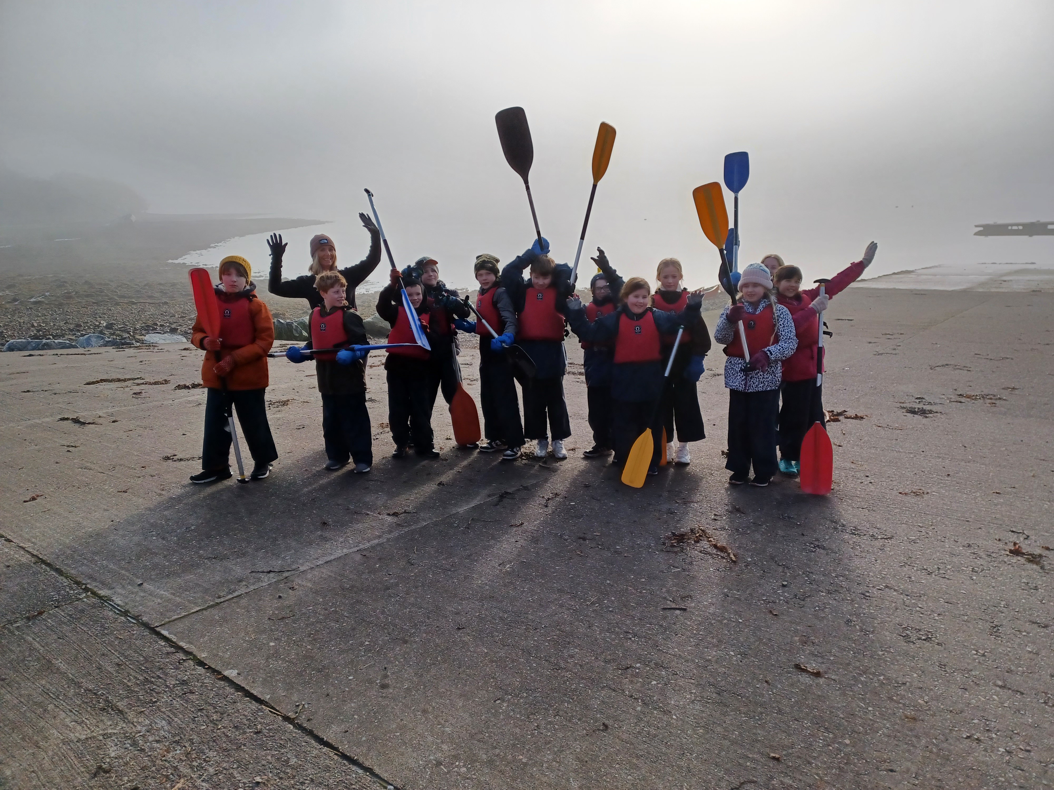 A group of children with kayak paddles standing on a misty beach looking very happy in waterproofs and some holding their paddles in the air.