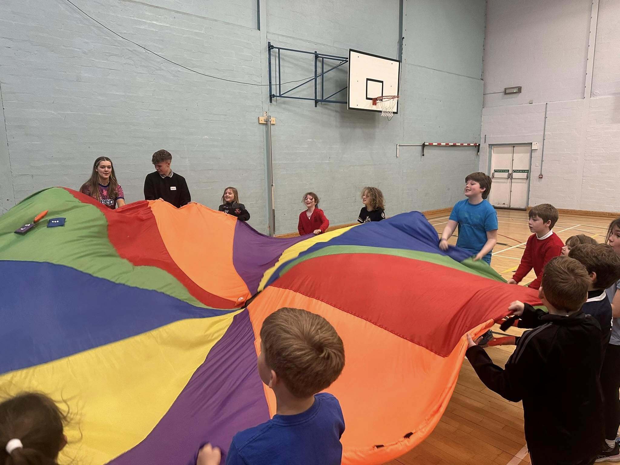 Smiling children gripping a colourful parachute in circular formation, waving it to bounce objects.