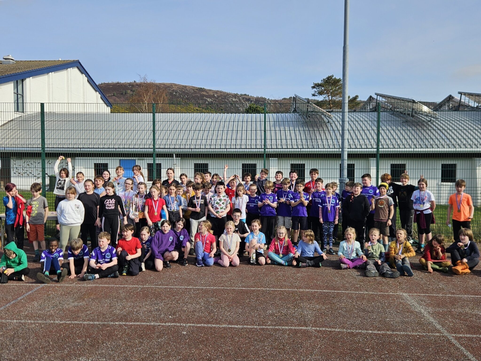 A large group of children posing on the edge of an outdoor pitch. Some have medals around their necks.