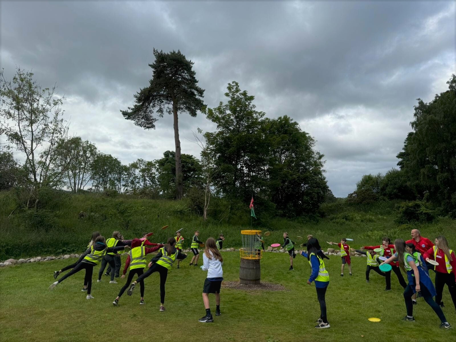 Many children playing disc golf in a field surrounded by trees. They are all throwing discs into a central target.