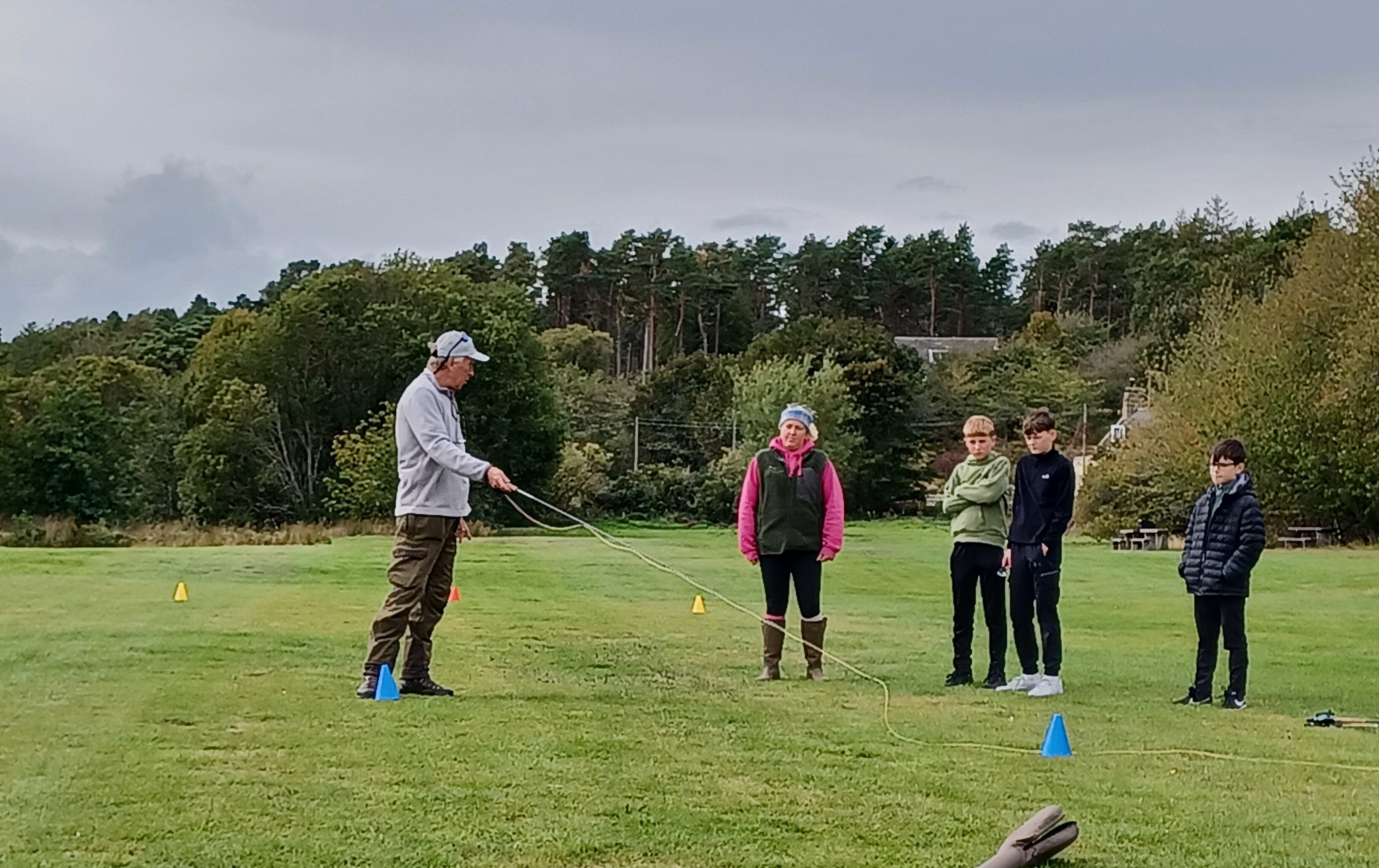 Man casting fly fishing rod in a field, with volunteer and three participants observing.