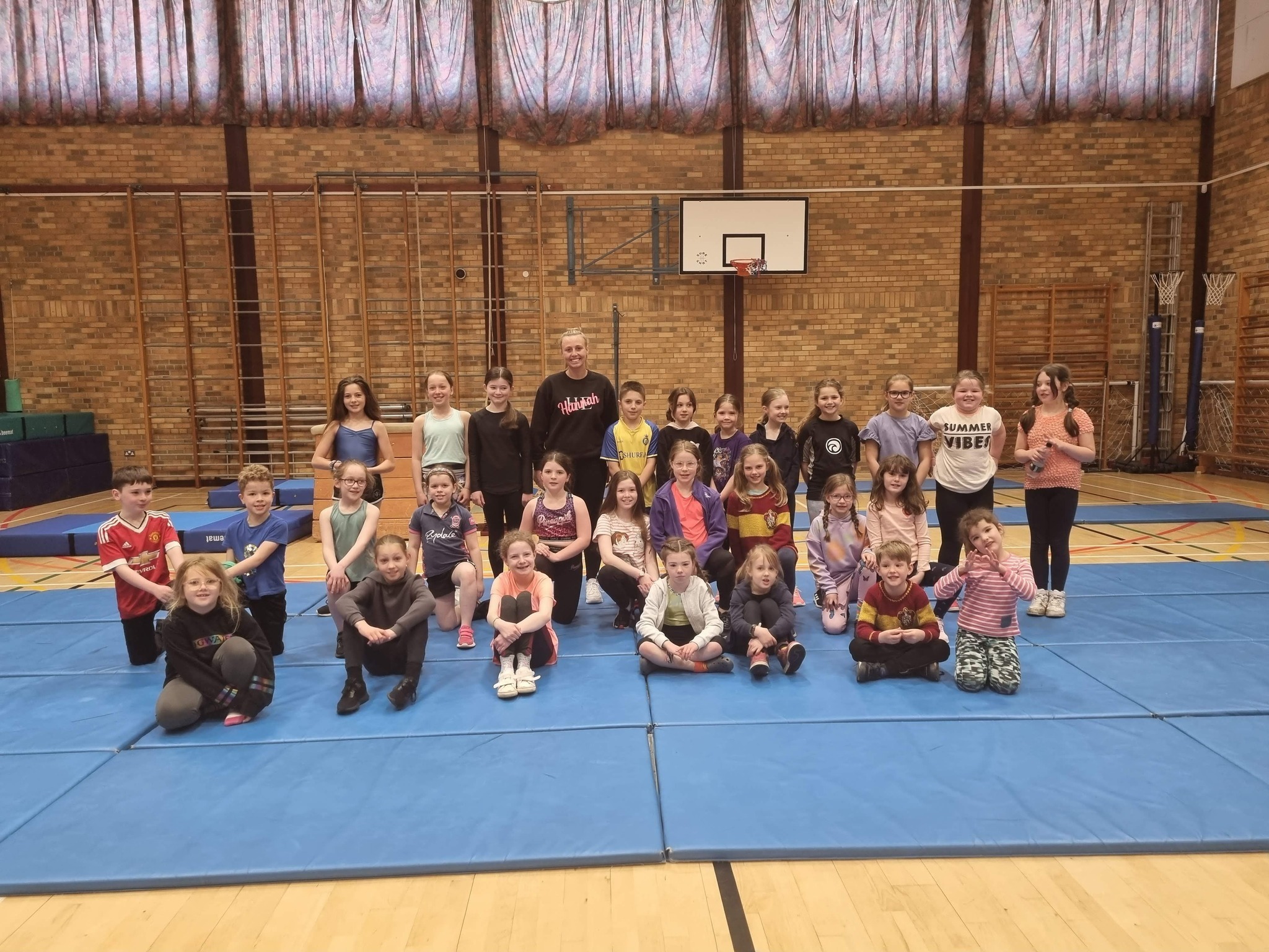 A large group of children sitting and standing on gym mats in a gym hall at gymnastics session.