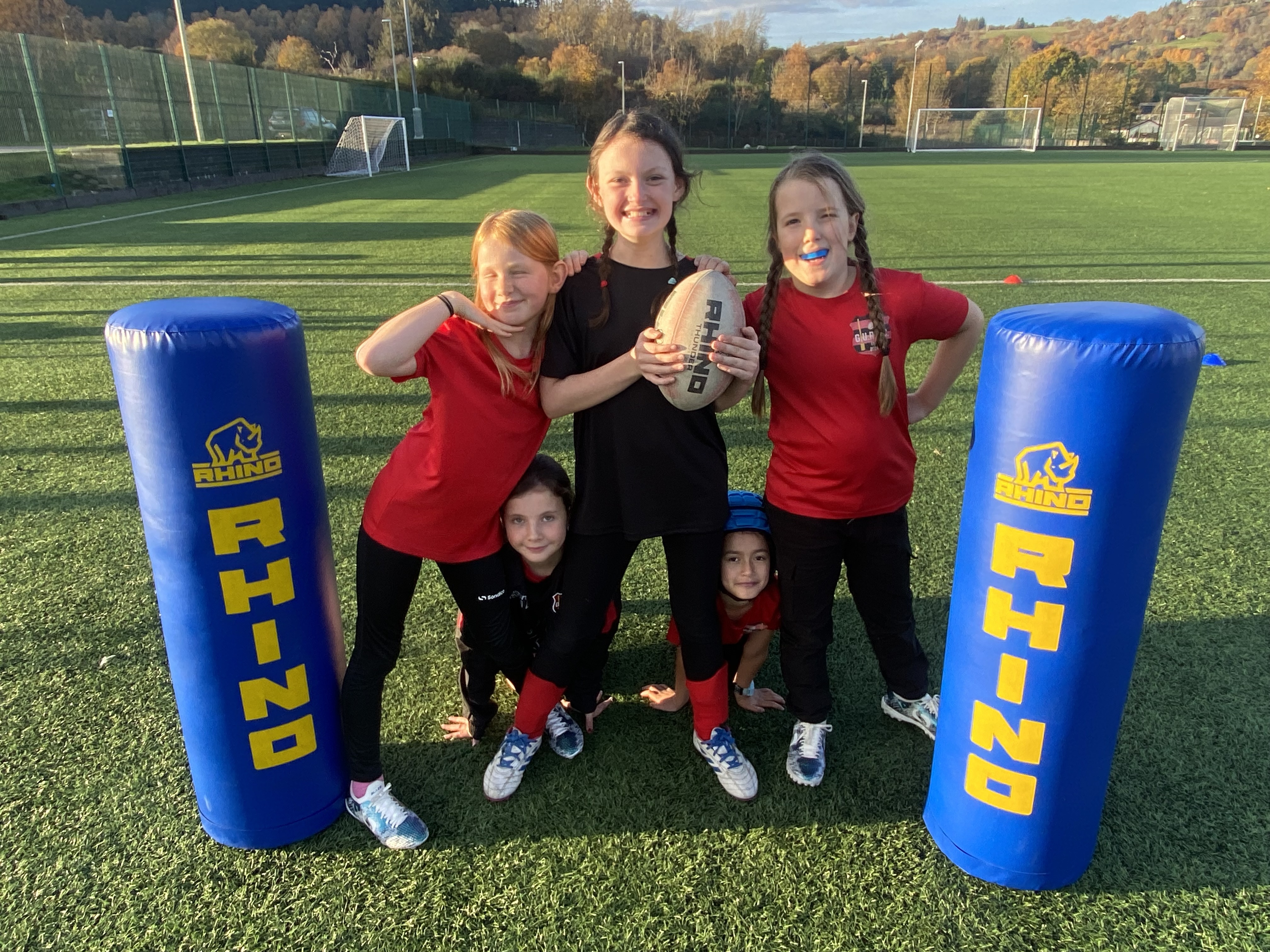 Five primary aged girls from Great Glen Cluster posing with rugby ball and rugby equipment.