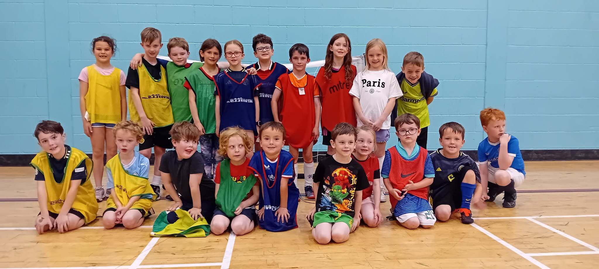 A group of young children sitting and standing at the edge of a gym hall wearing different colours bibs.