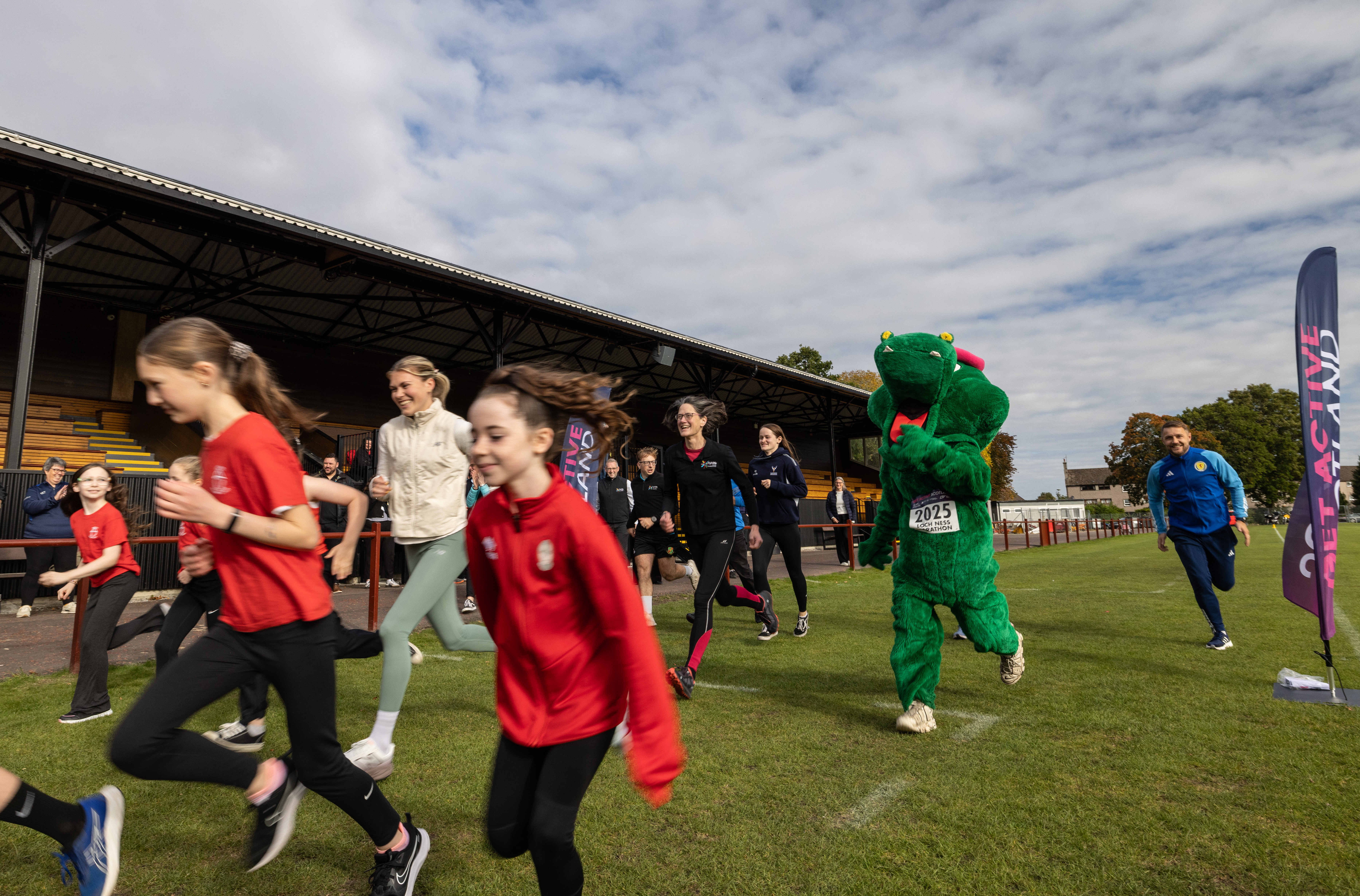 Children running on a sports field alongside a person in a green mascot costume.