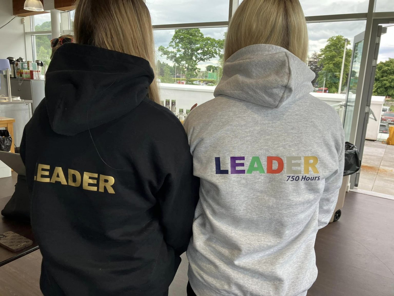 Two people standing indoors wearing hoodies with the word ‘LEADER’ printed on the back, one in black and one in grey with multicolored text.
