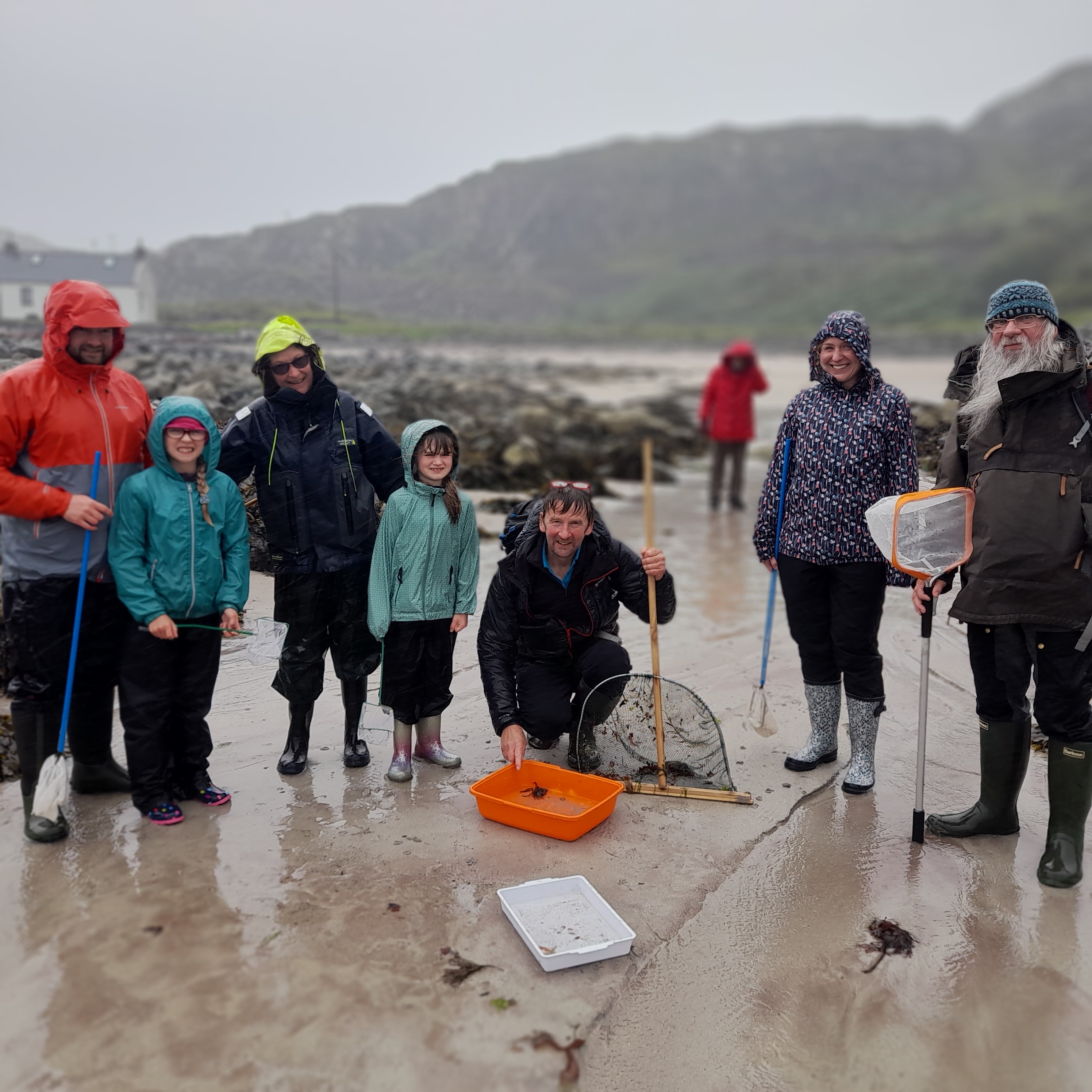 Group of people on a sandy beach during a rainy day, wearing waterproof jackets and boots. They are gathered around trays and nets used for exploring marine life, with rocky shoreline and hills visible in the background.