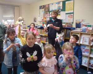 Group of children standing in a library holding paper crafts shaped like insects, with bookshelves and posters visible in the background. An adult is assisting with the activity.
