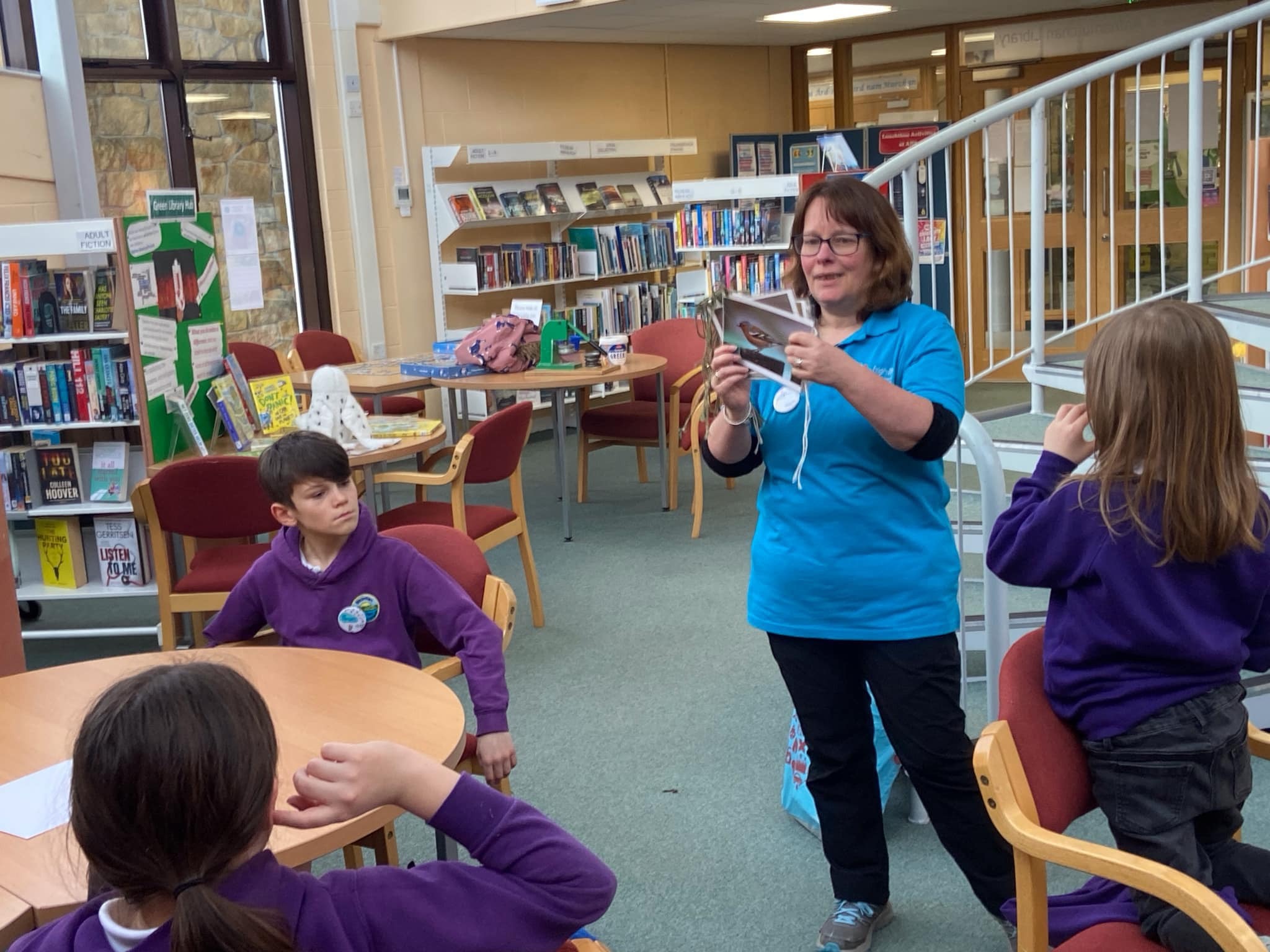 Person in a blue polo shirt showing a picture card to children seated around tables in a library with bookshelves and a staircase.