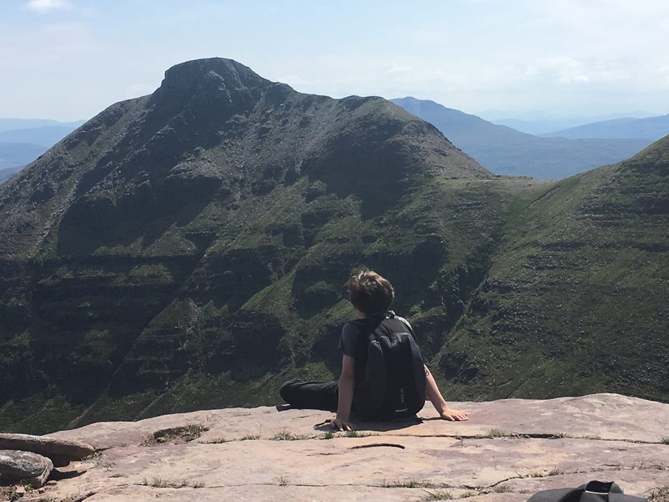 A person sitting on a stone slab on the top of a hill looking at the view of a ridge with a clear sky above them