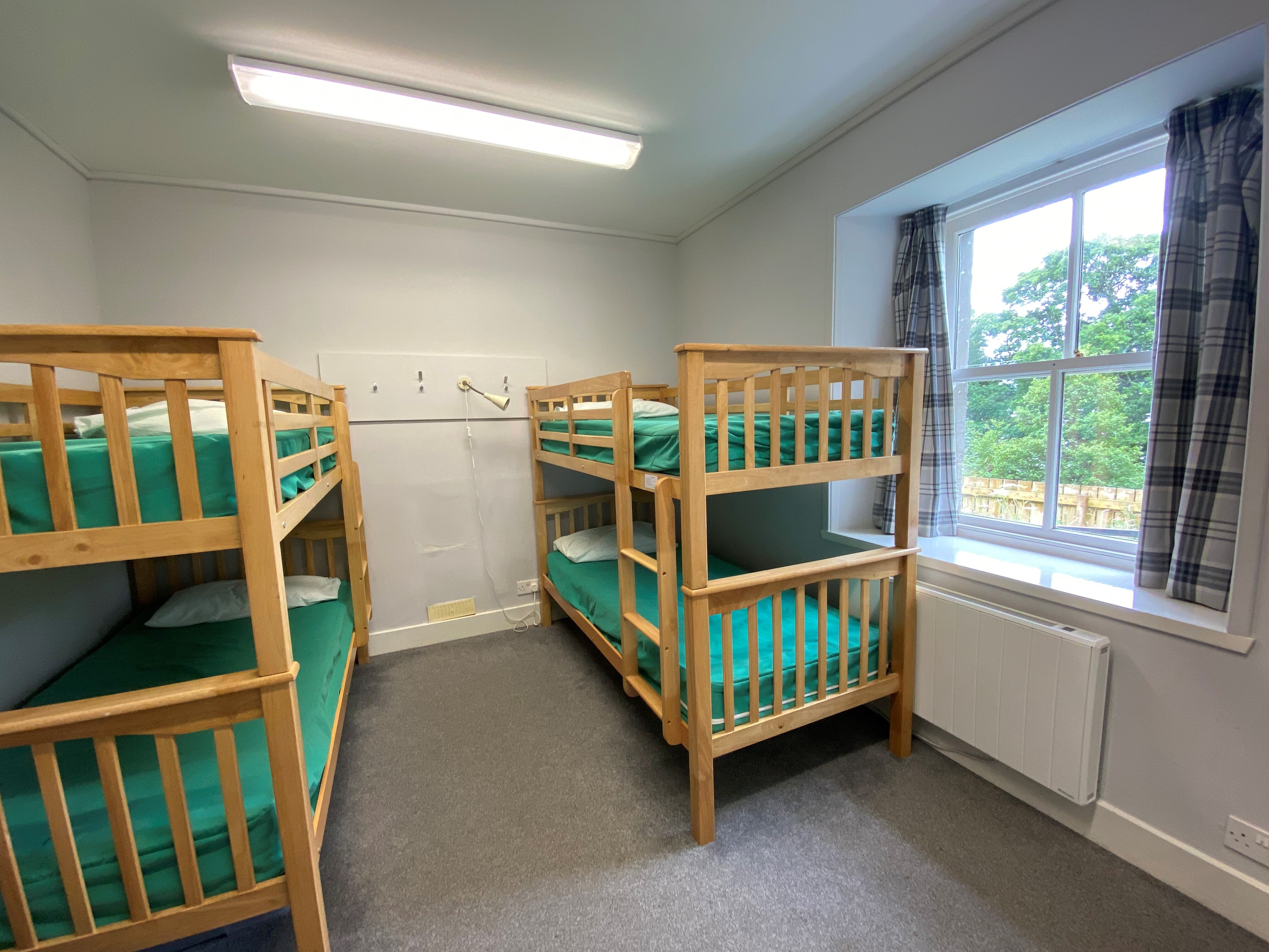 Room with two wooden bunk beds fitted with green bedding, a window with plaid curtains, grey carpeted floor, and white walls.