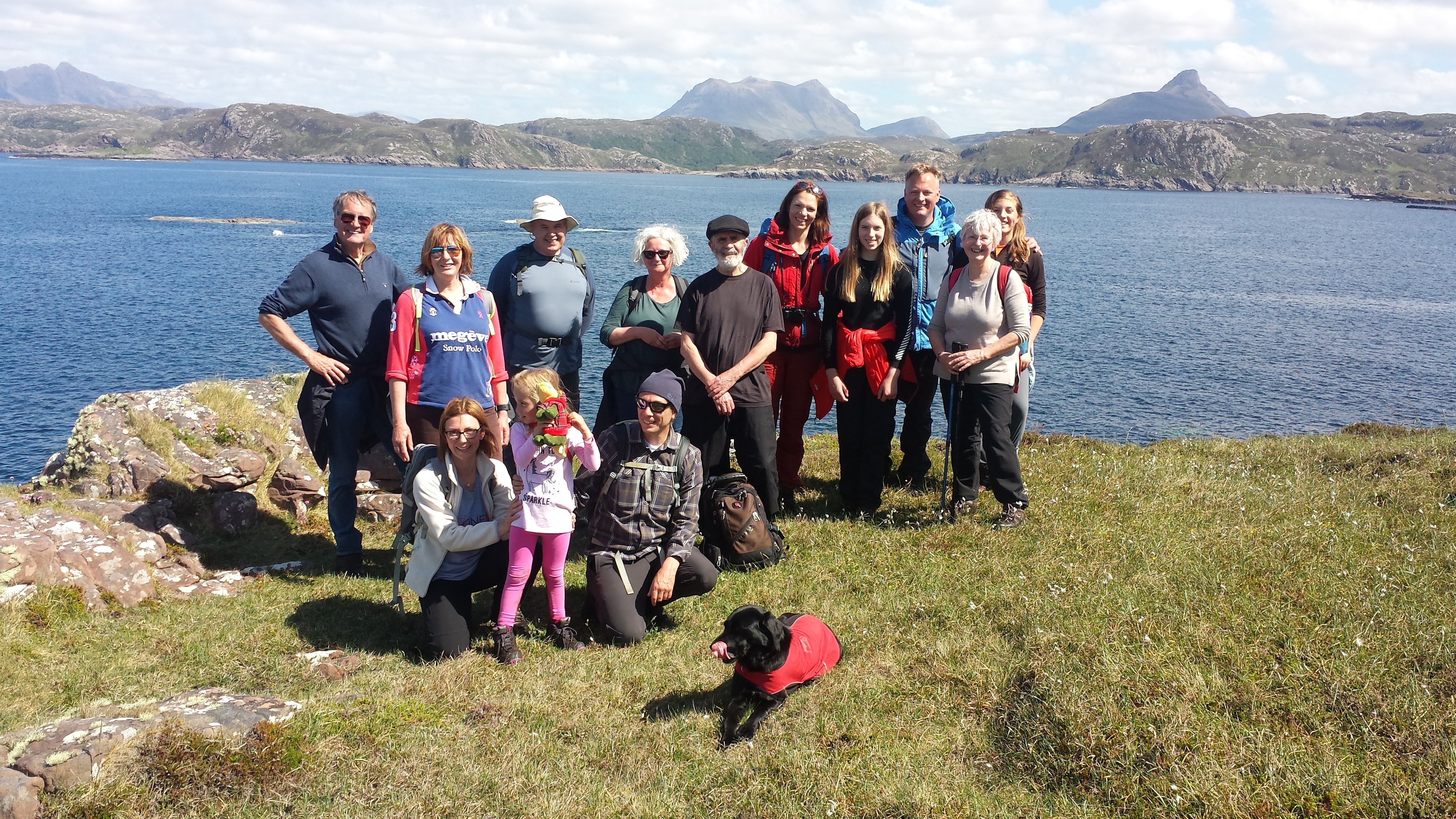 An intergenerational group of around 15 people standing by the sea, on a clear day with a view of mountains in the distance across the water