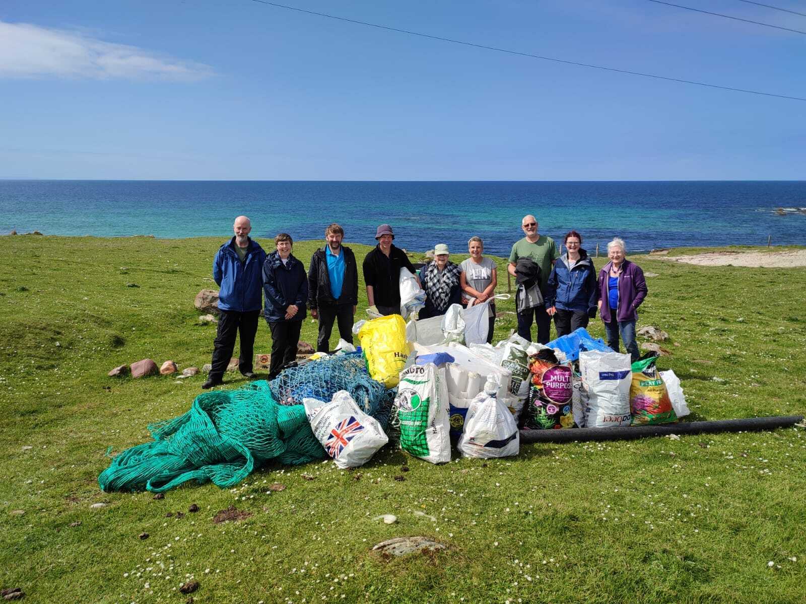Group of volunteers standing on grassy ground near the sea with piles of collected rubbish and fishing nets in bags.
