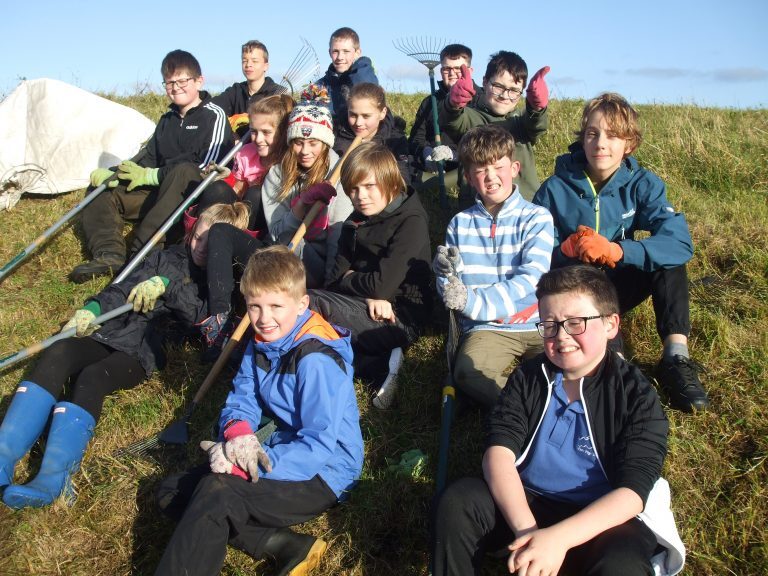 Group of young volunteers sitting on a grassy slope holding rakes and wearing gloves, gathered for a conservation task under clear blue skies.