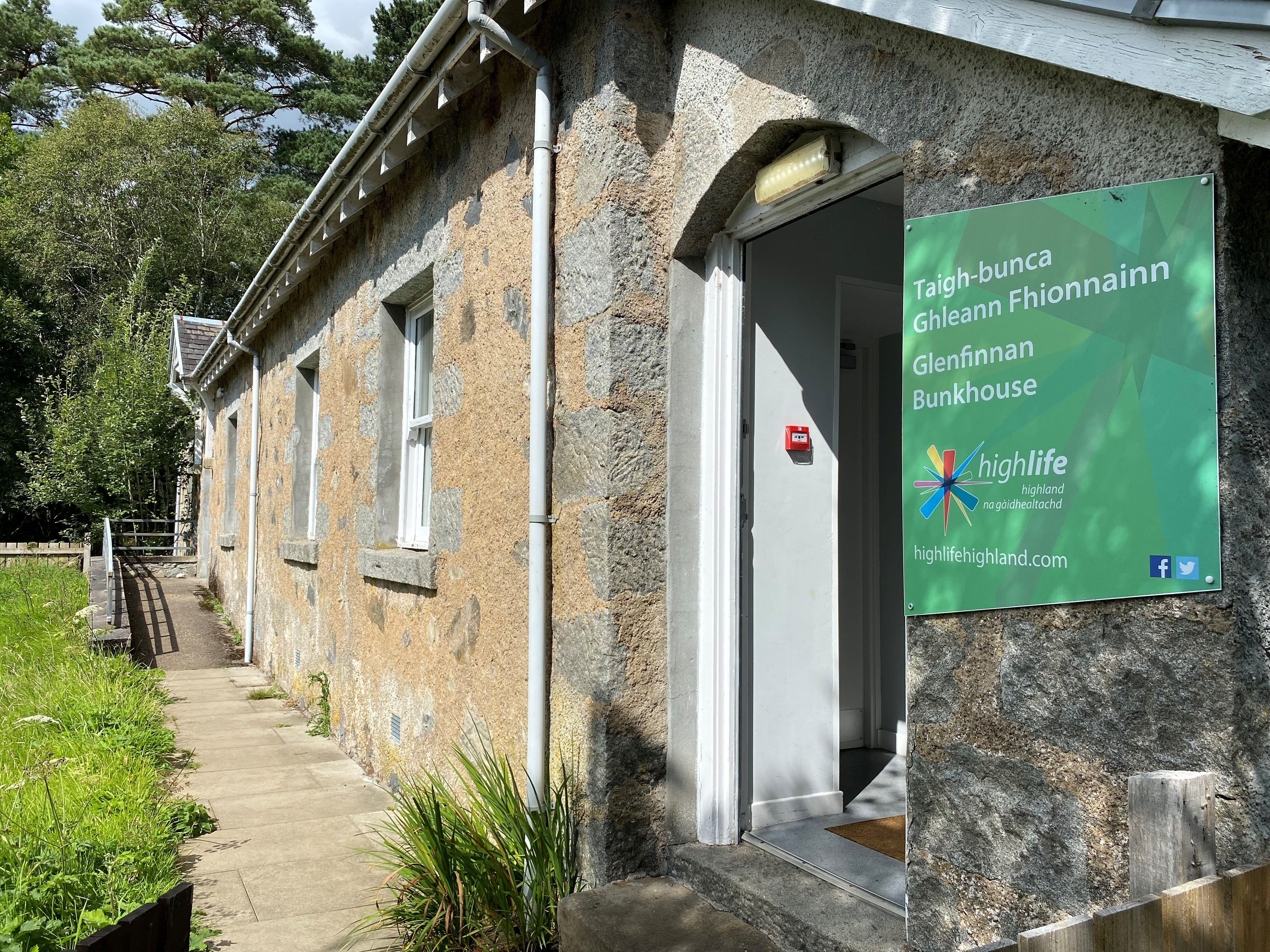 A stone building with a green sign reading “Glenfinnan Bunkhouse” beside an open doorway, surrounded by trees and greenery on a sunny day.