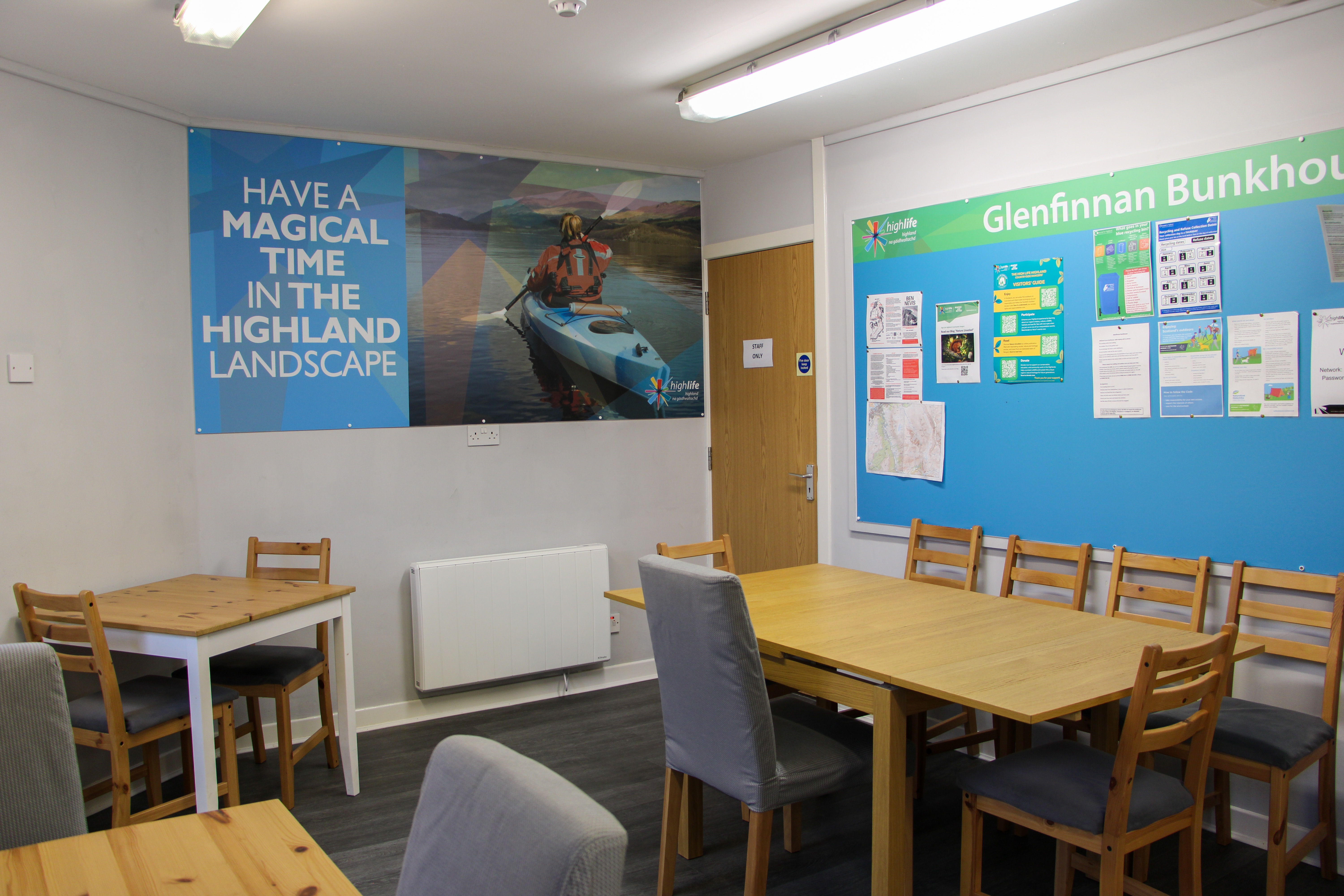 Dining room with wooden tables and mixed chairs, a blue noticeboard labeled “Glenfinnan Bunkhouse,” and a large wall poster showing a kayaker with the text “Have a magical time in the Highland landscape.”