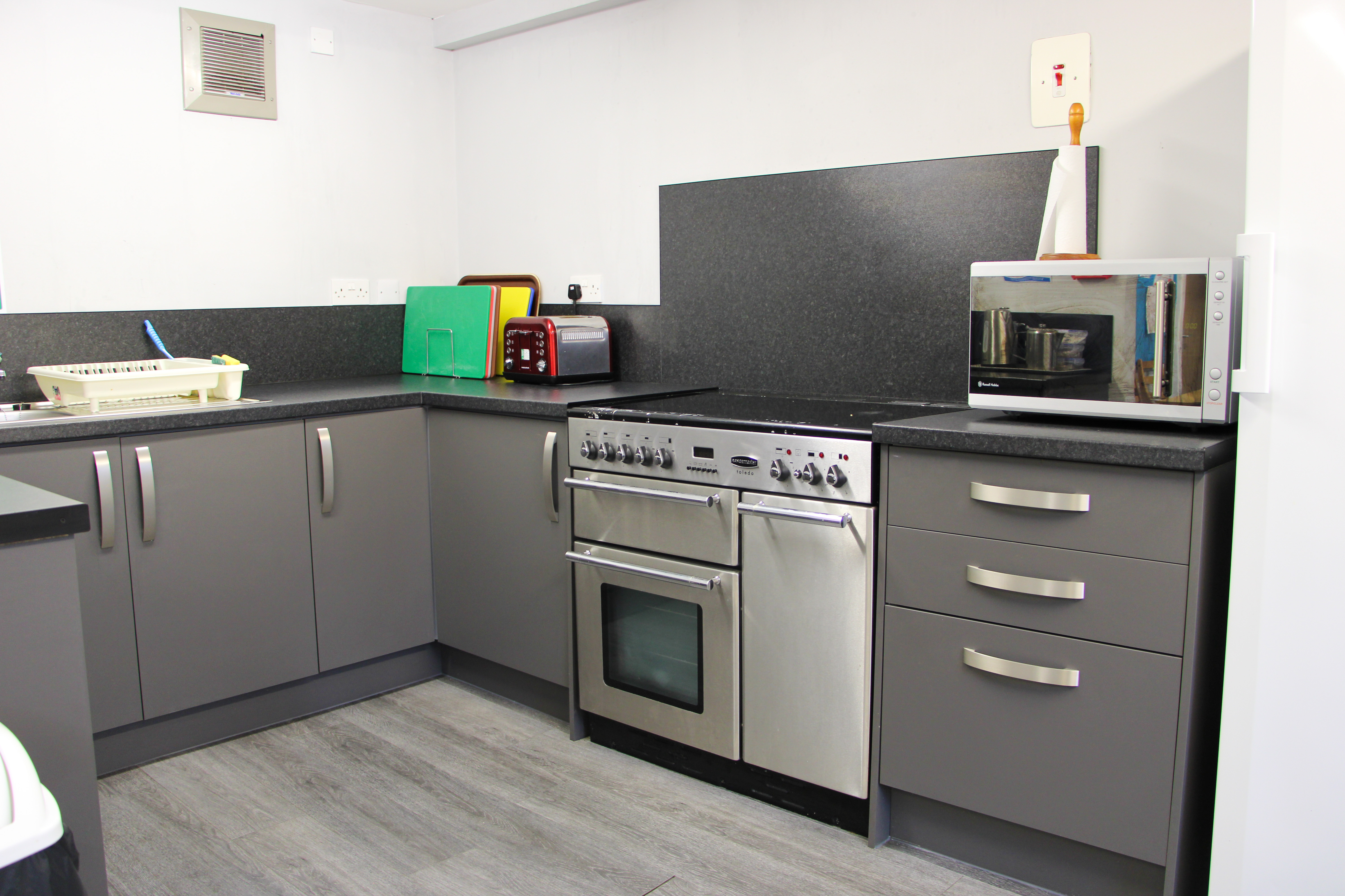 A modern kitchen with grey cabinets, a stainless steel oven, black countertops, and appliances including a microwave, toaster, and dish rack.