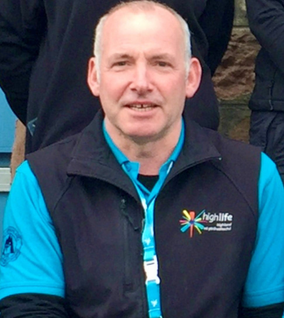 Countryside Ranger wearing a blue High Life Highland polo shirt and black fleece vest with logo, seated in front of a stone wall.