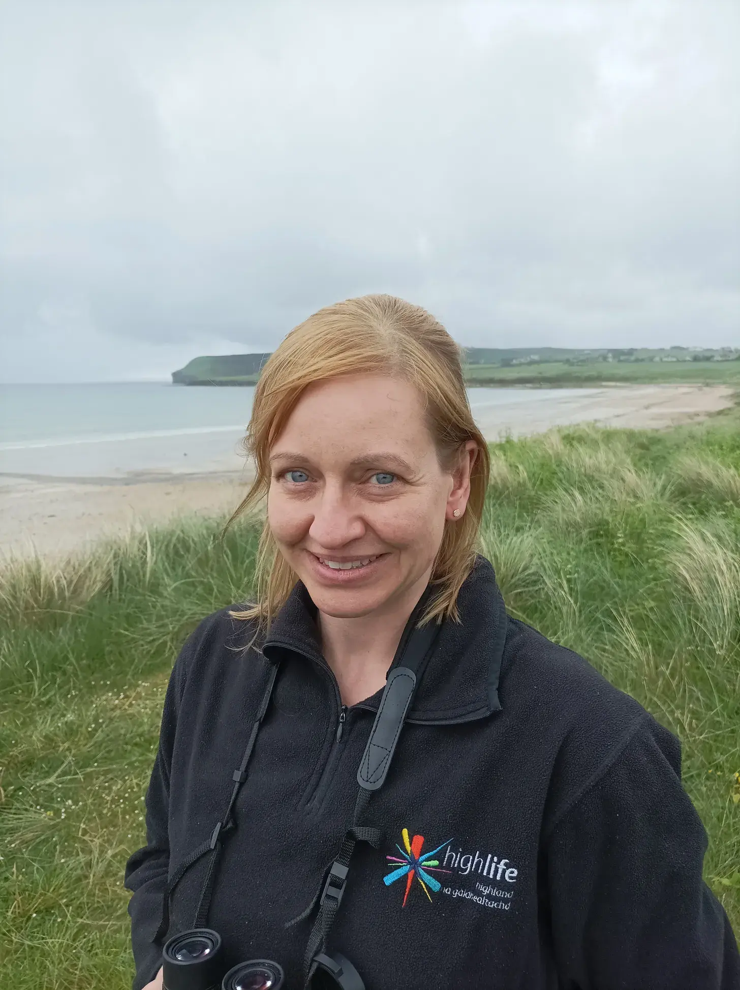 Countryside Ranger in a black High Life Highland fleece holding binoculars on a coastal grassy area with sea and cliffs behind.