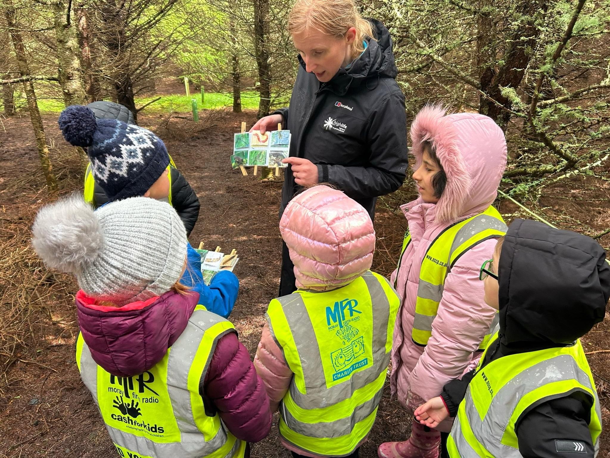 A female ranger is standing in a woodland with 5 children showing them a nature chart