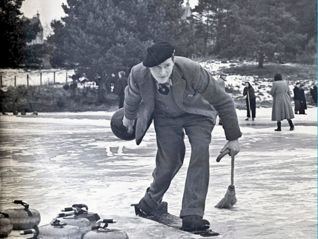 Person playing curling on an outdoor frozen surface, holding a curling stone and broom, with several stones on the ice and snow-covered trees in the background.