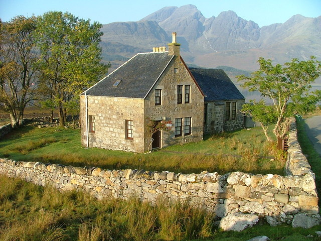 Stone-built house with a slate roof and chimneys, surrounded by a low stone wall, grassy ground, and a few trees, set against a backdrop of tall, rugged mountains in soft sunlight.