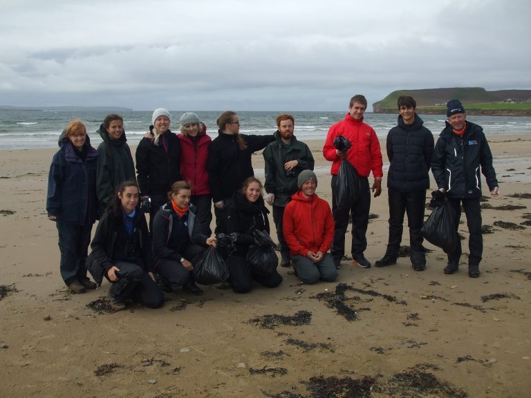 Group of volunteers standing on a sandy beach holding black rubbish bags after a coastal clean-up, with the sea and distant headland in the background.
