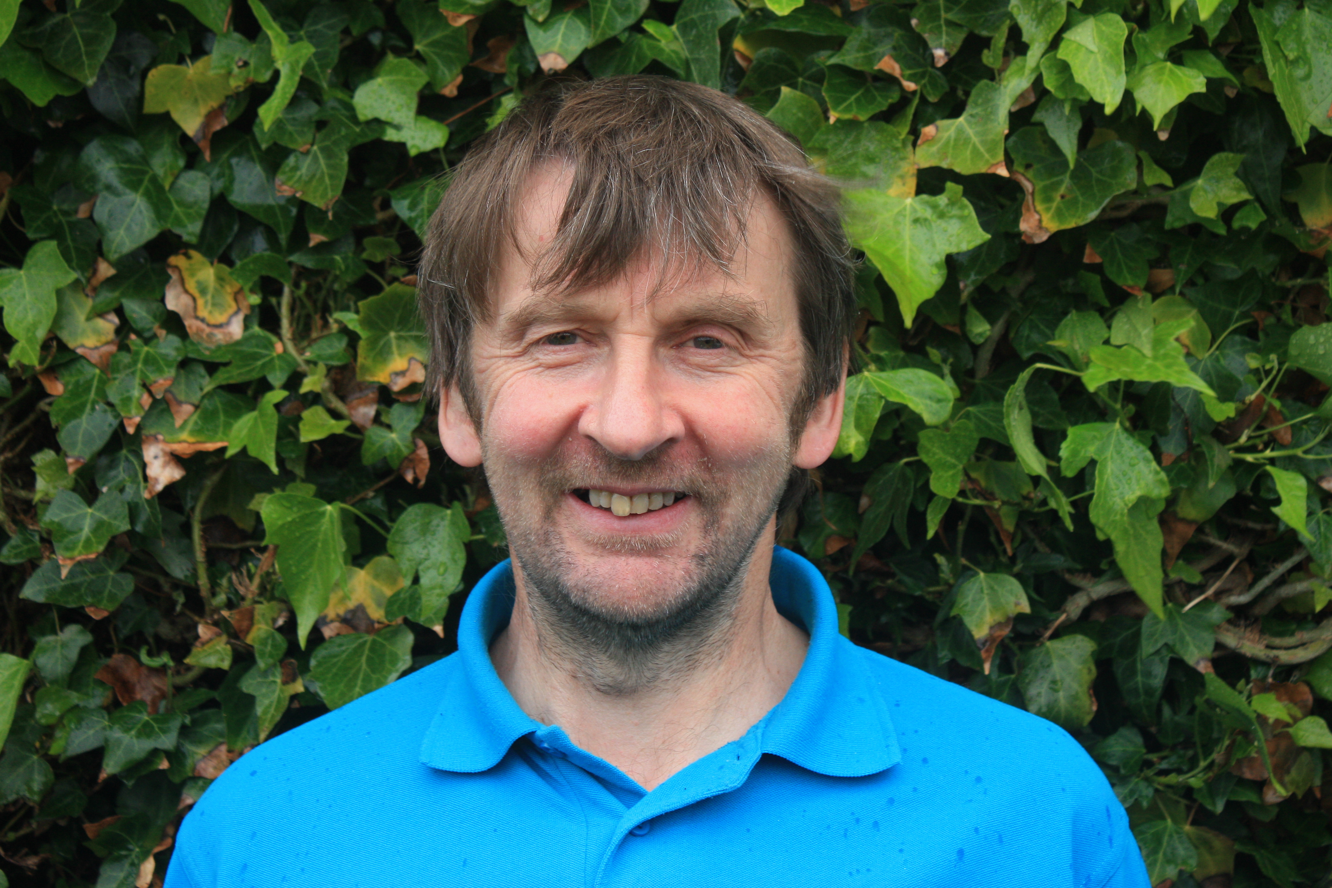 Countryside Ranger in a blue High Life Highland polo shirt standing outdoors in front of a wall of green ivy.