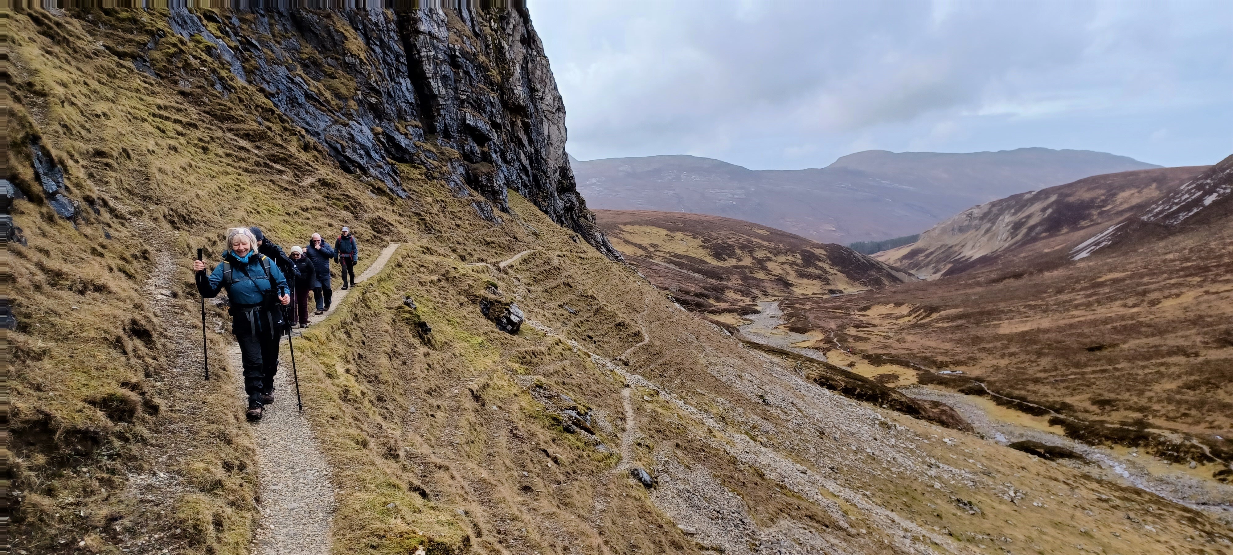 Group of walkers on a narrow path along a steep rocky hillside in a remote valley with mountains in the distance.