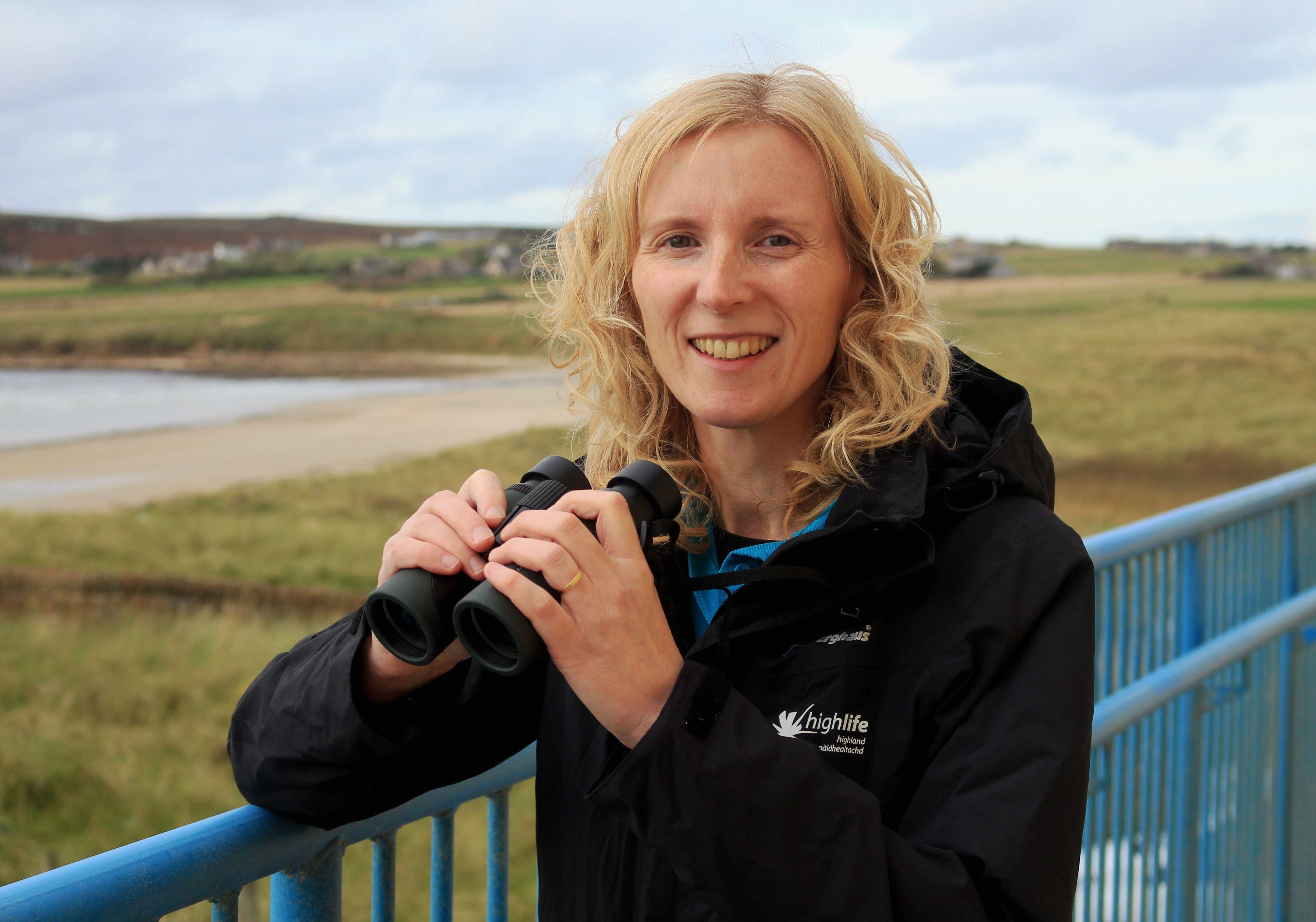 Countryside Ranger in a black High Life Highland jacket holding binoculars on a bridge overlooking a sandy beach and grassy area.