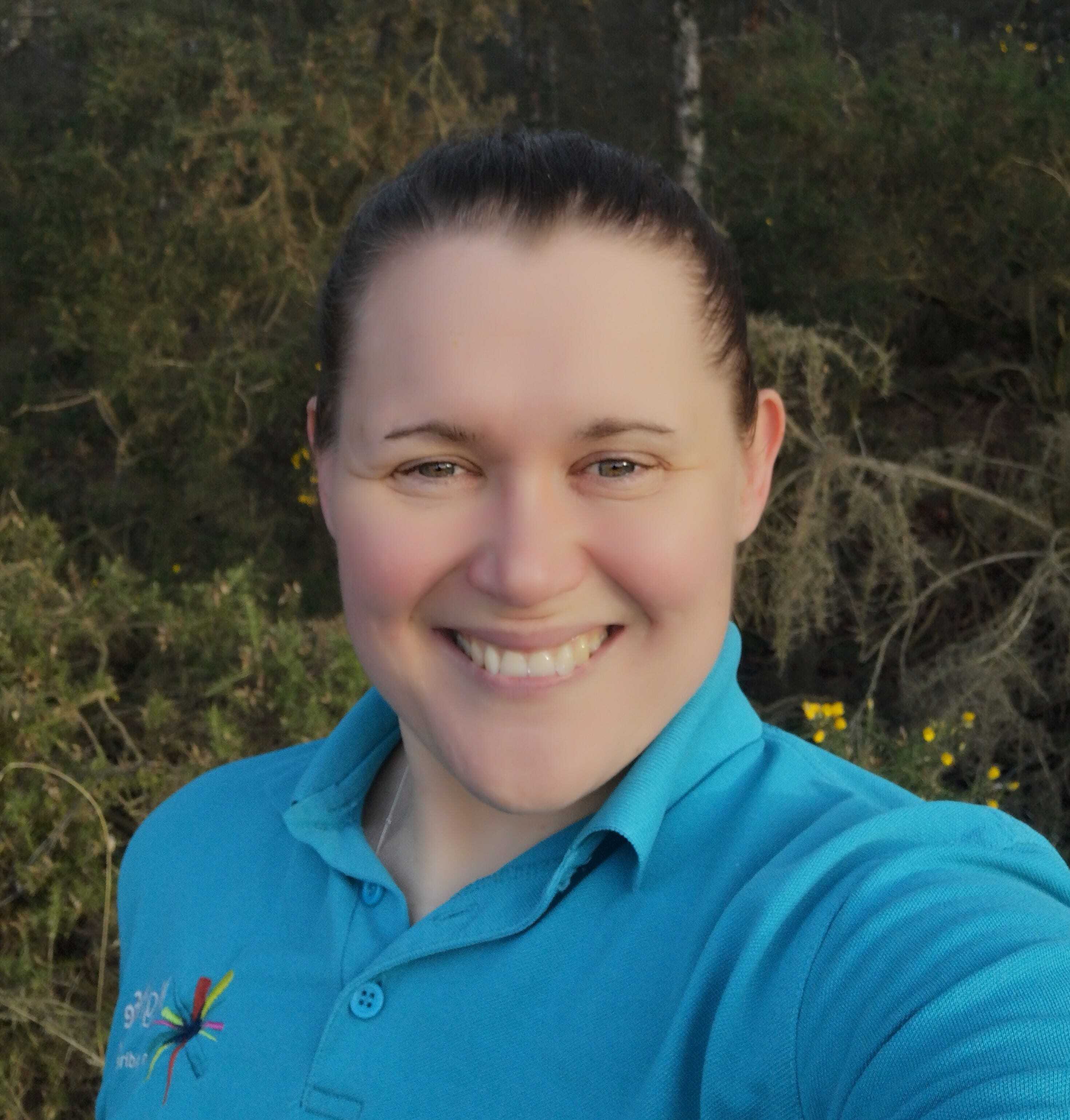 Countryside Ranger in a blue High Life Highland polo shirt outdoors with trees and shrubs in the background.