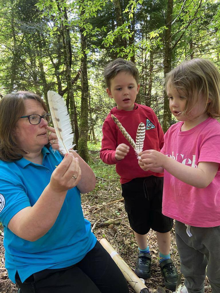 Person in a blue polo shirt showing a large white feather to two children holding smaller feathers in a woodland setting.