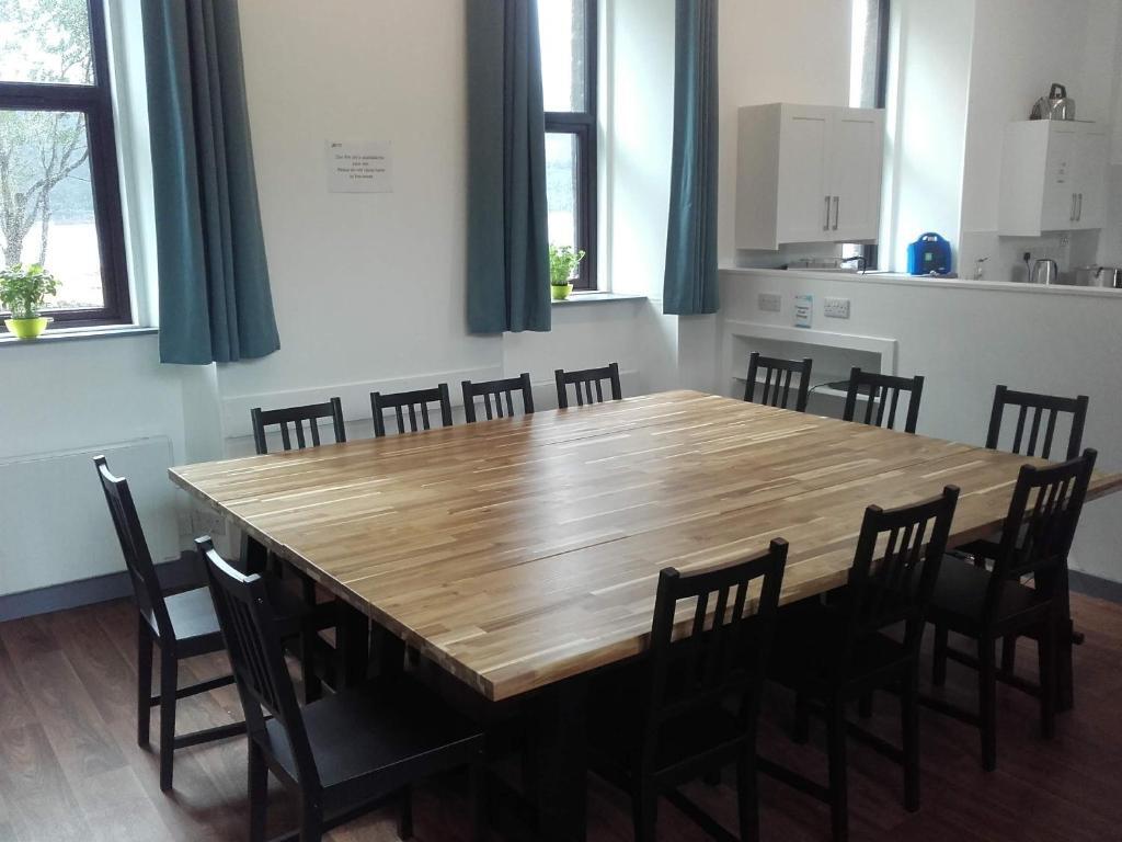 Communal dining area with a large wooden table surrounded by black chairs, set in a bright room with windows and white walls.