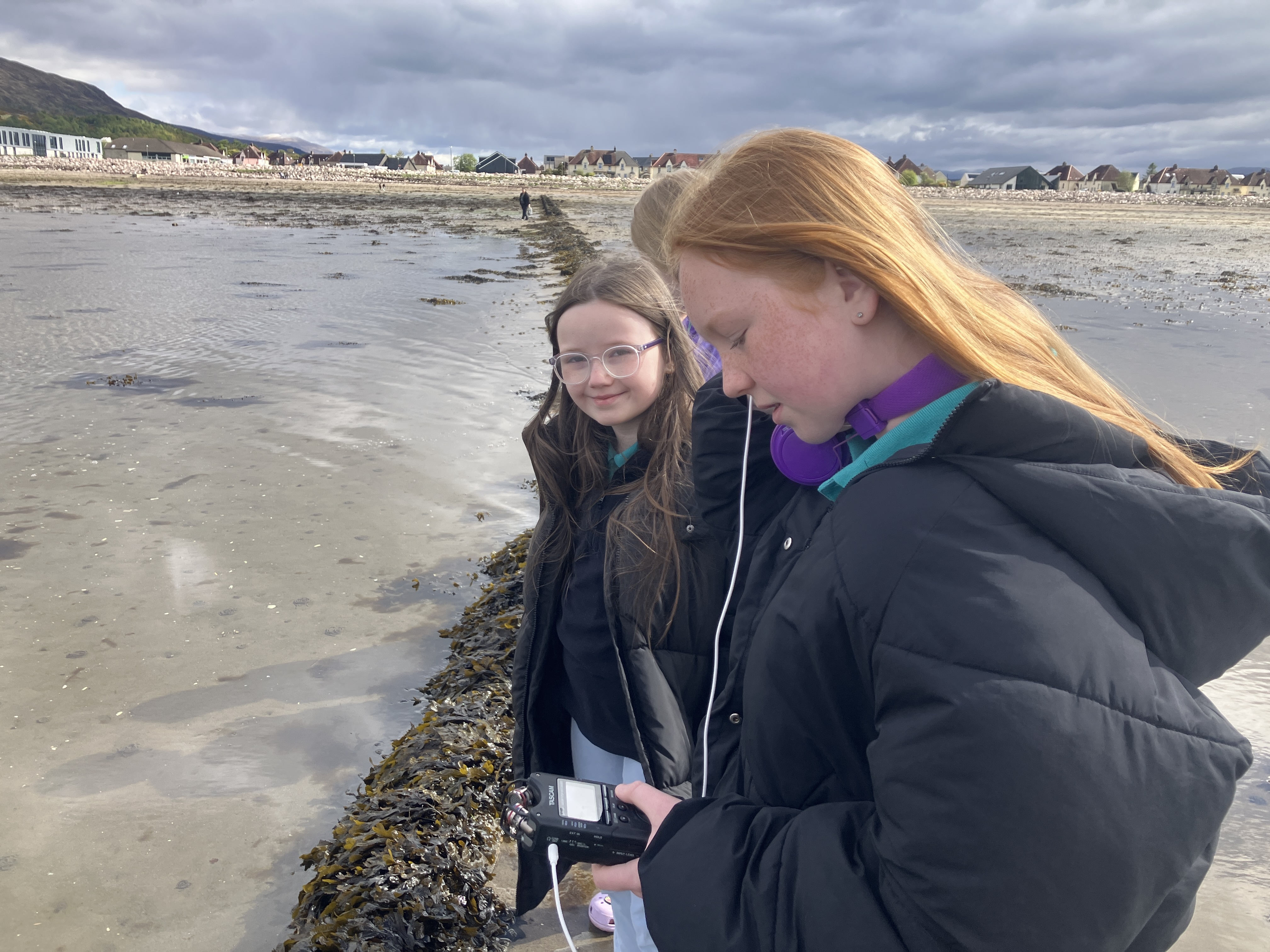 Two primary school pupils on the shoreline. They are both wearing black jackets. One has long dark hair and one has long red hair. The girl closest to the camera is holding sounds recording equipment. In the background there is a beach and houses in the distance.